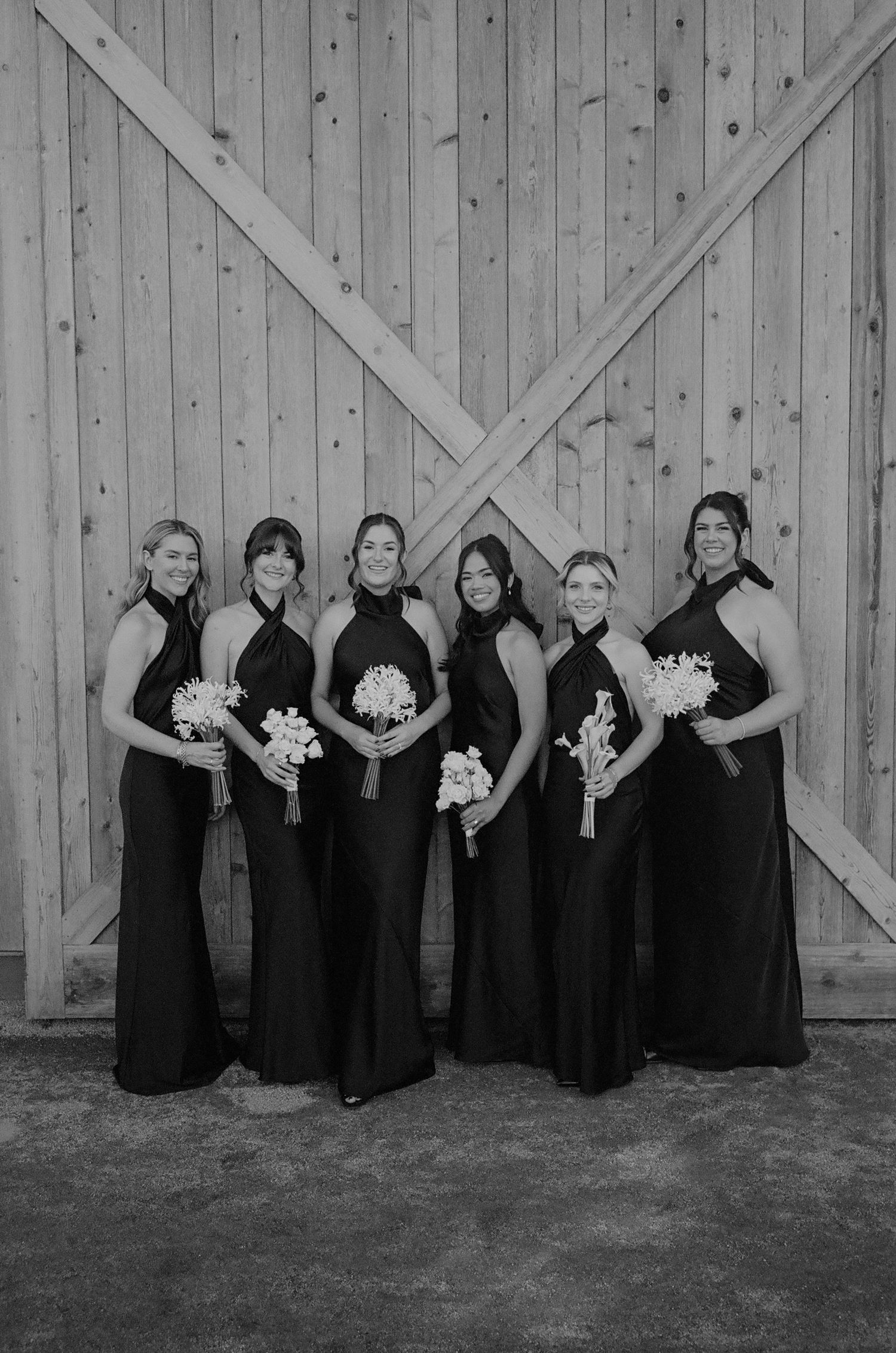 Bridesmaids in elegant black dresses hold white bouquets while posing against a rustic wooden barn wall.
