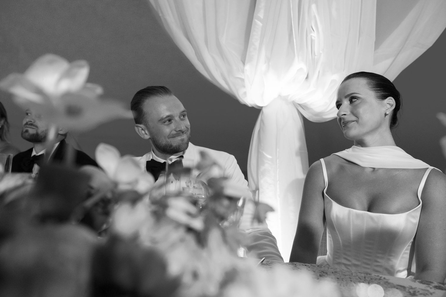 A couple shares an intimate moment against white fabric in black and white.