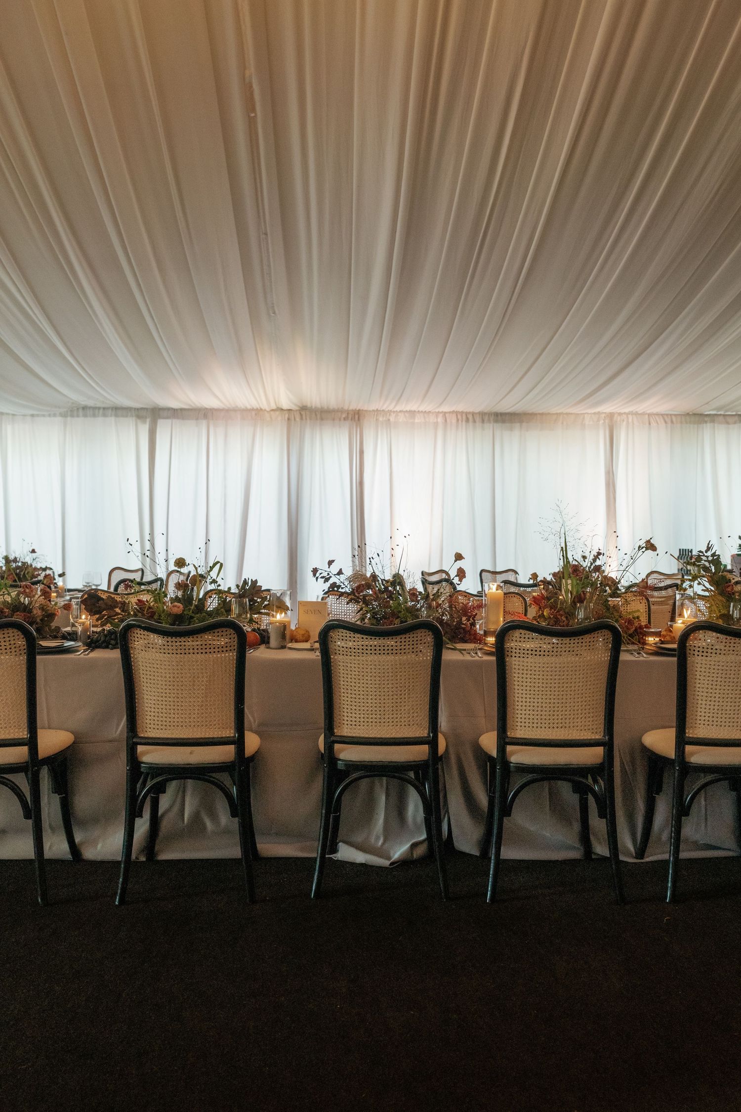 Long banquet tables with cane-backed chairs arranged in rows under white tent draping for an elegant reception.