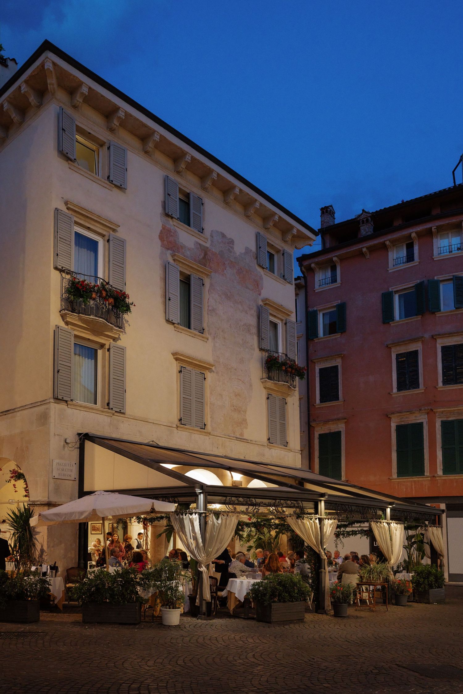 Outdoor restaurant terrace lit at dusk in an Italian piazza, surrounded by historic colorful buildings.