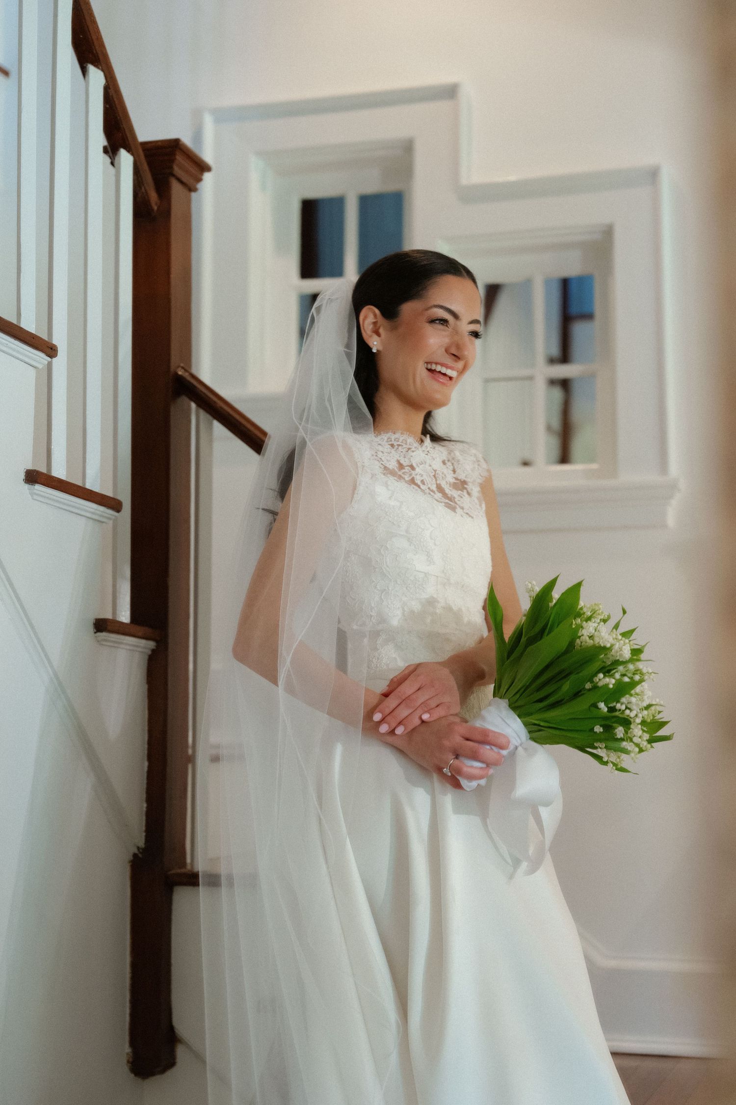 Smiling bride in white lace gown and veil holds lily of the valley bouquet on elegant staircase.
