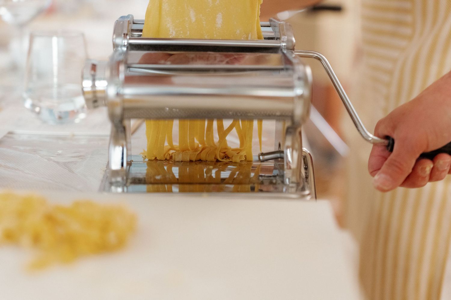Fresh pasta being cut through a stainless steel pasta machine on a white marble surface.
