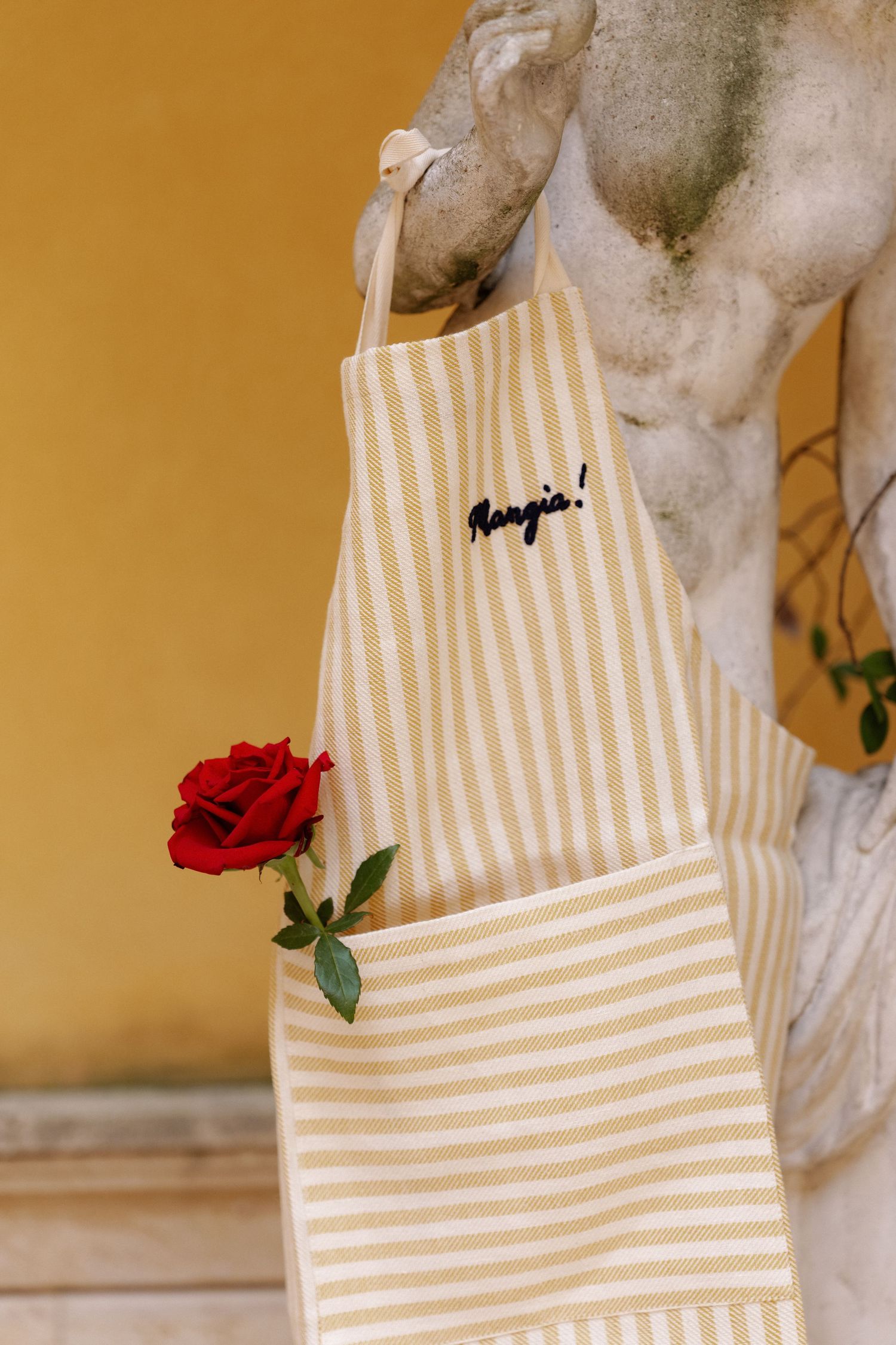 Yellow striped tote bag with Mangia embroidery and red rose, hanging on classical stone sculpture.