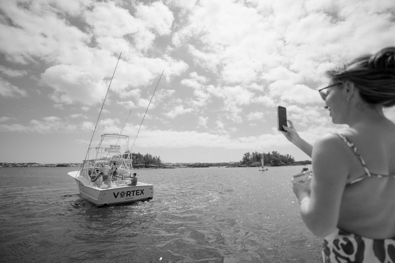 A sailboat is seen on the water with a person standing on the shore taking a photograph.