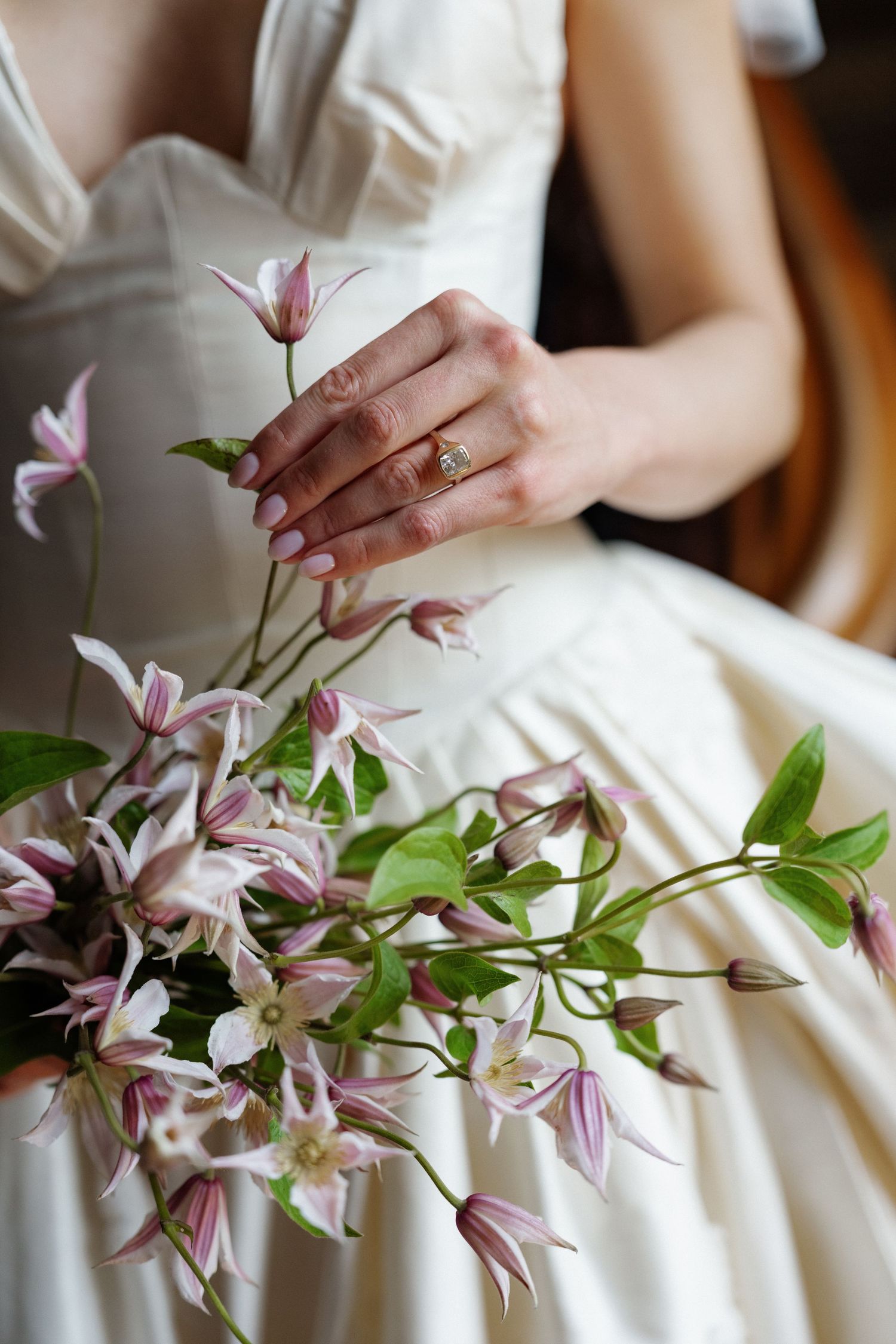 Bride in white gown holding delicate pink clematis flowers, wearing a gold engagement ring on her manicured hand.