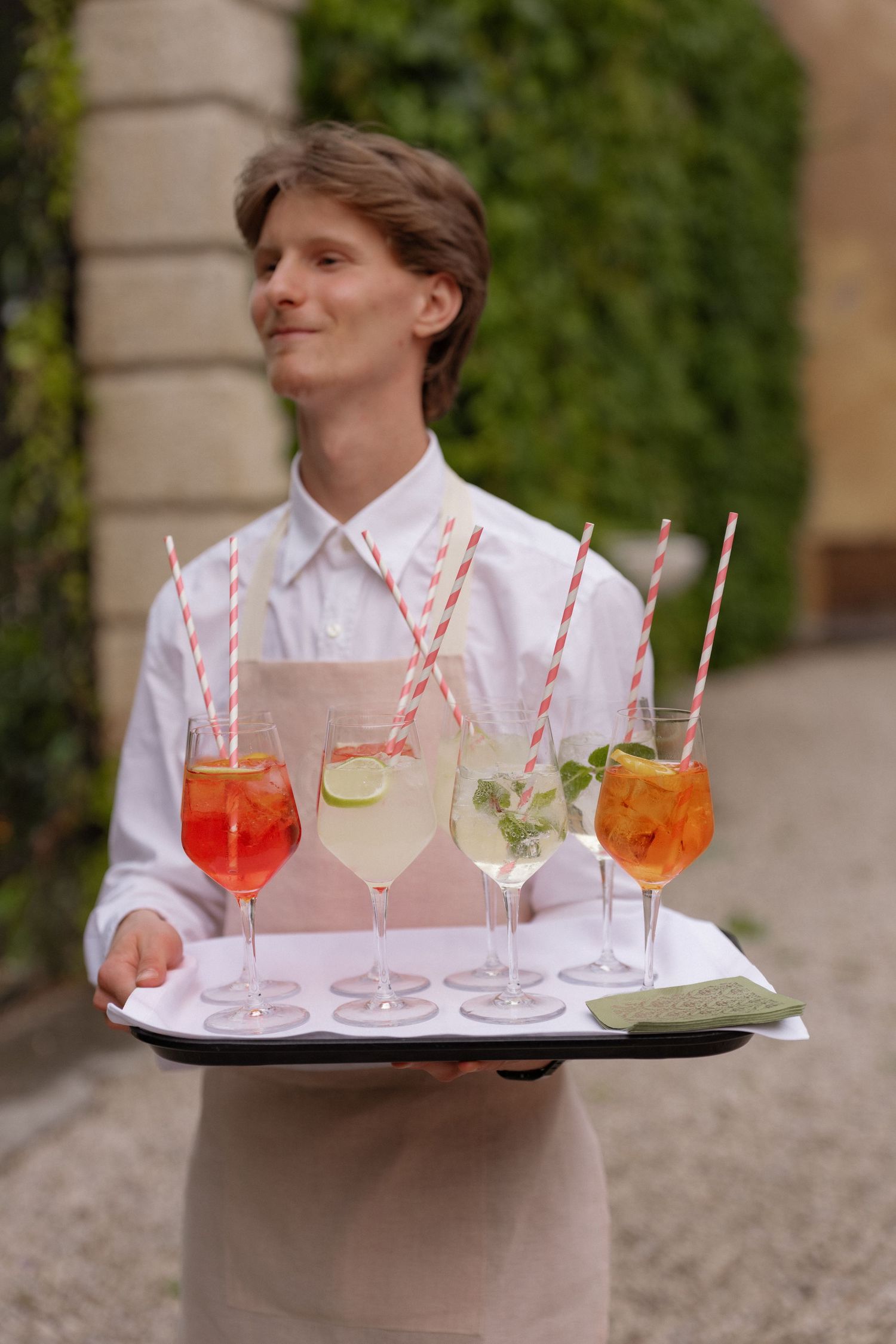Smiling waiter holding a tray of colorful cocktails with striped straws in an outdoor garden setting.