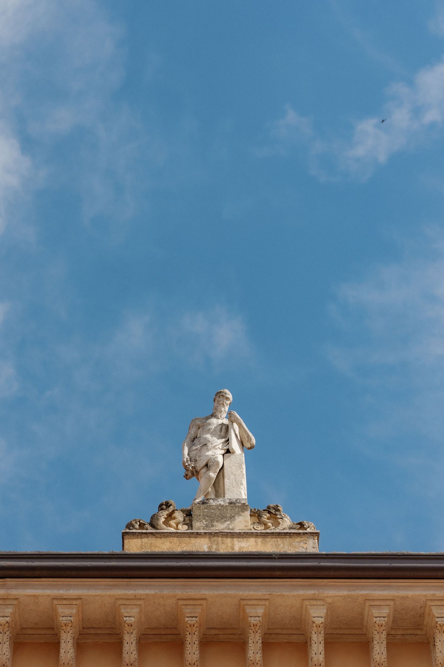 Classical stone statue atop a neoclassical building facade against a blue sky with light clouds.