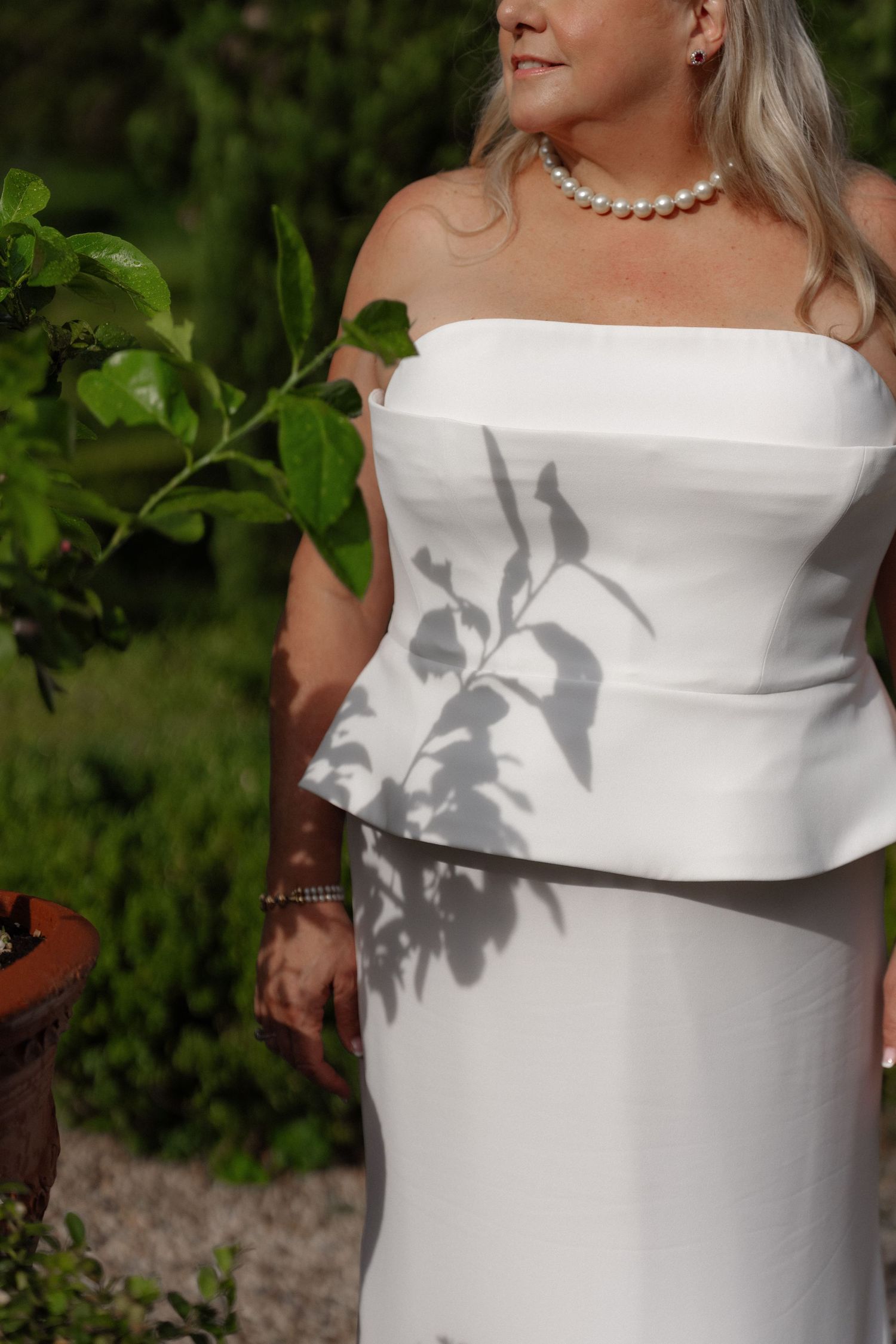 Bride in white strapless peplum gown with pearl necklace standing in a lush garden with leaf shadows.
