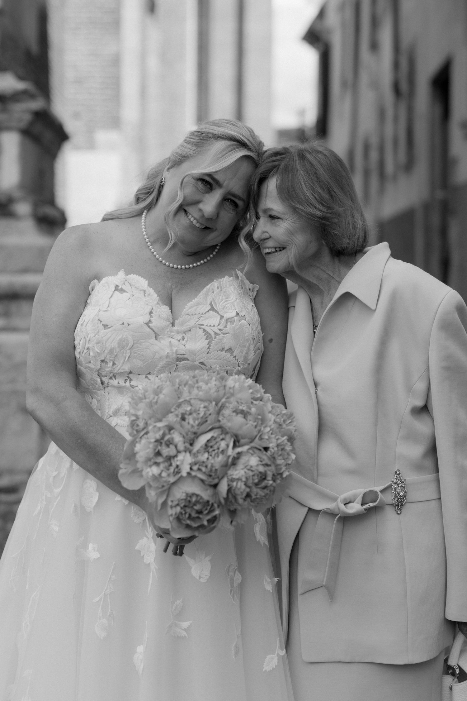 Black and white photo of a bride in lace gown holding floral bouquet posing with older woman in elegant suit.