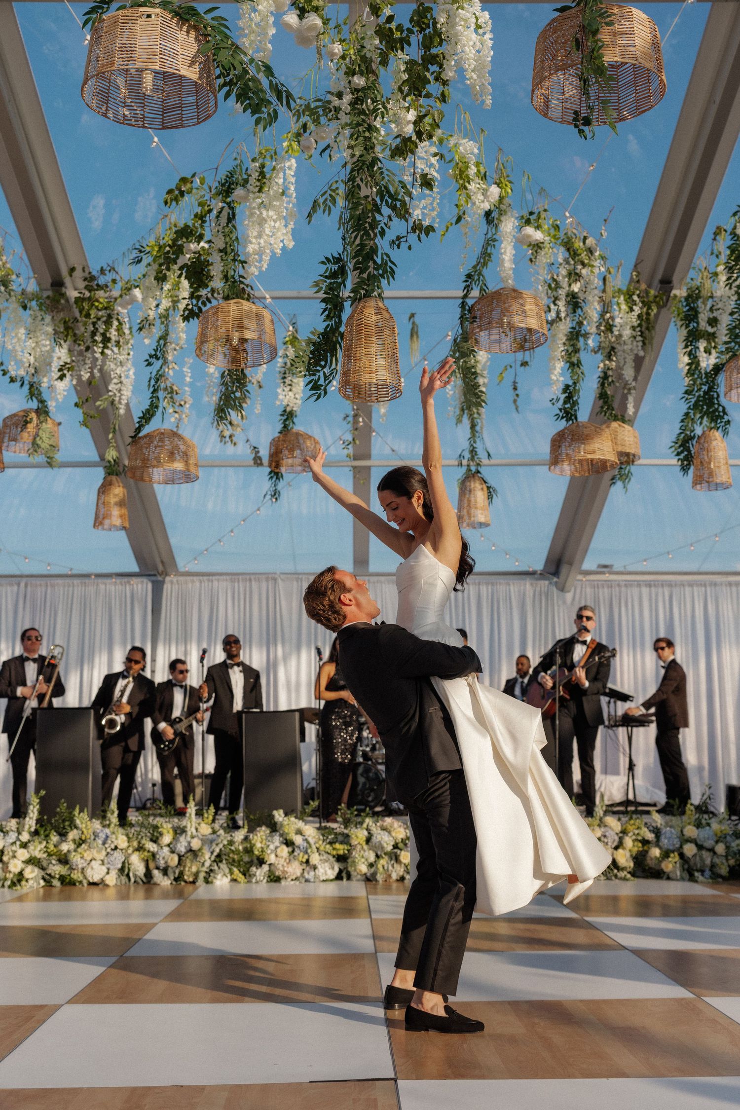 Bride and groom share first dance under clear tent with live band, greenery, and wicker lanterns.