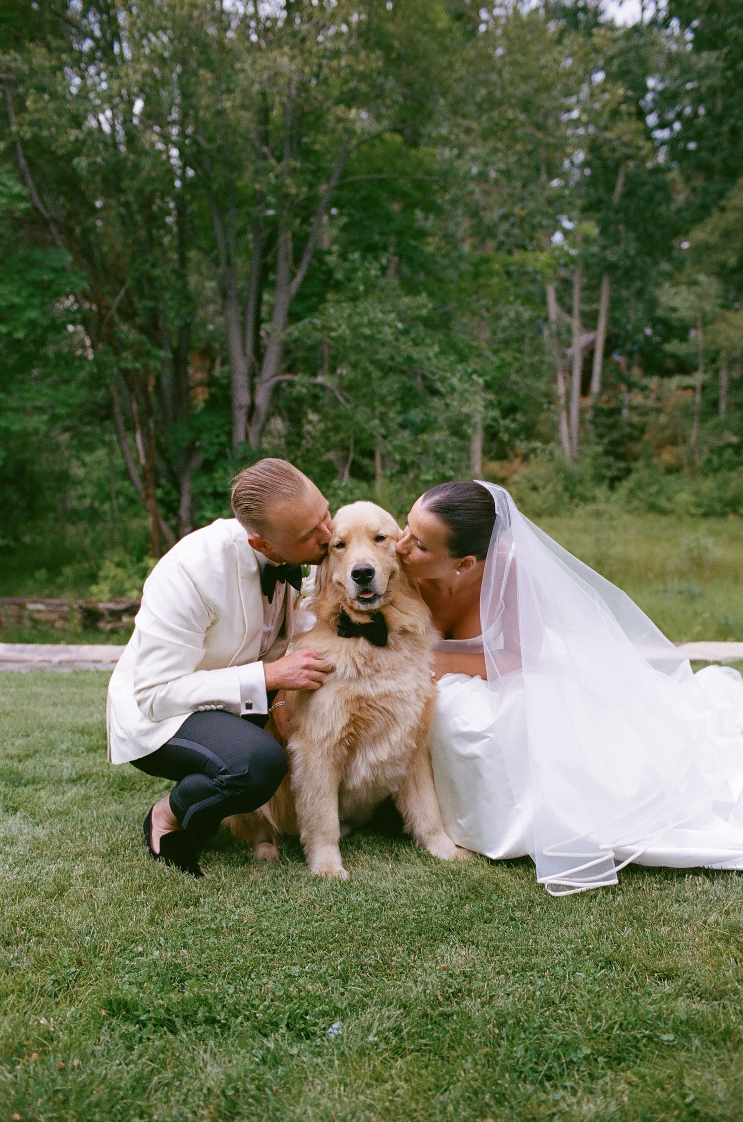 A happy golden retriever shares a tender moment with a wedding couple on a grassy lawn surrounded by trees.
