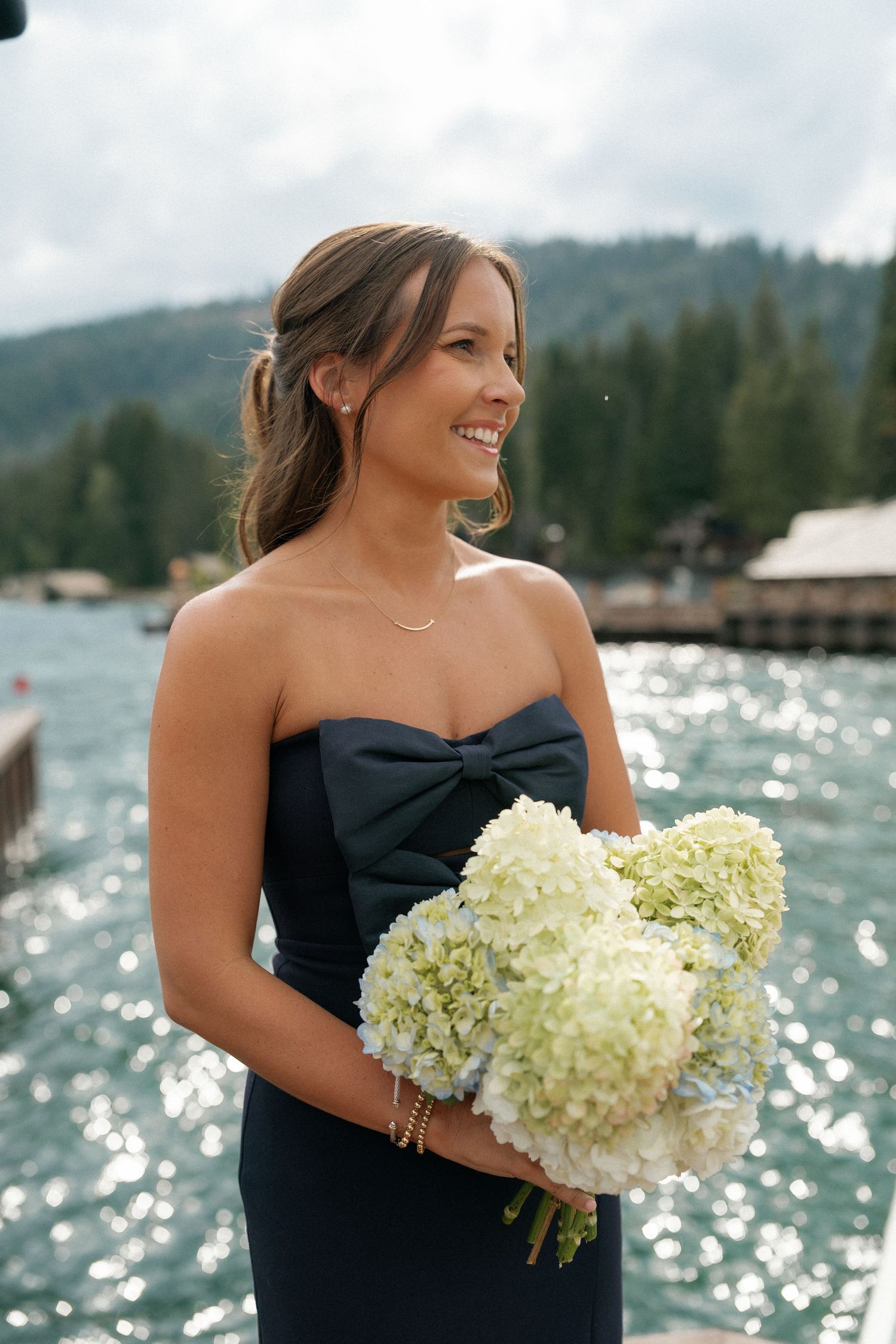 Wedding attendant in navy dress holding white hydrangea bouquet by sparkling waterfront.