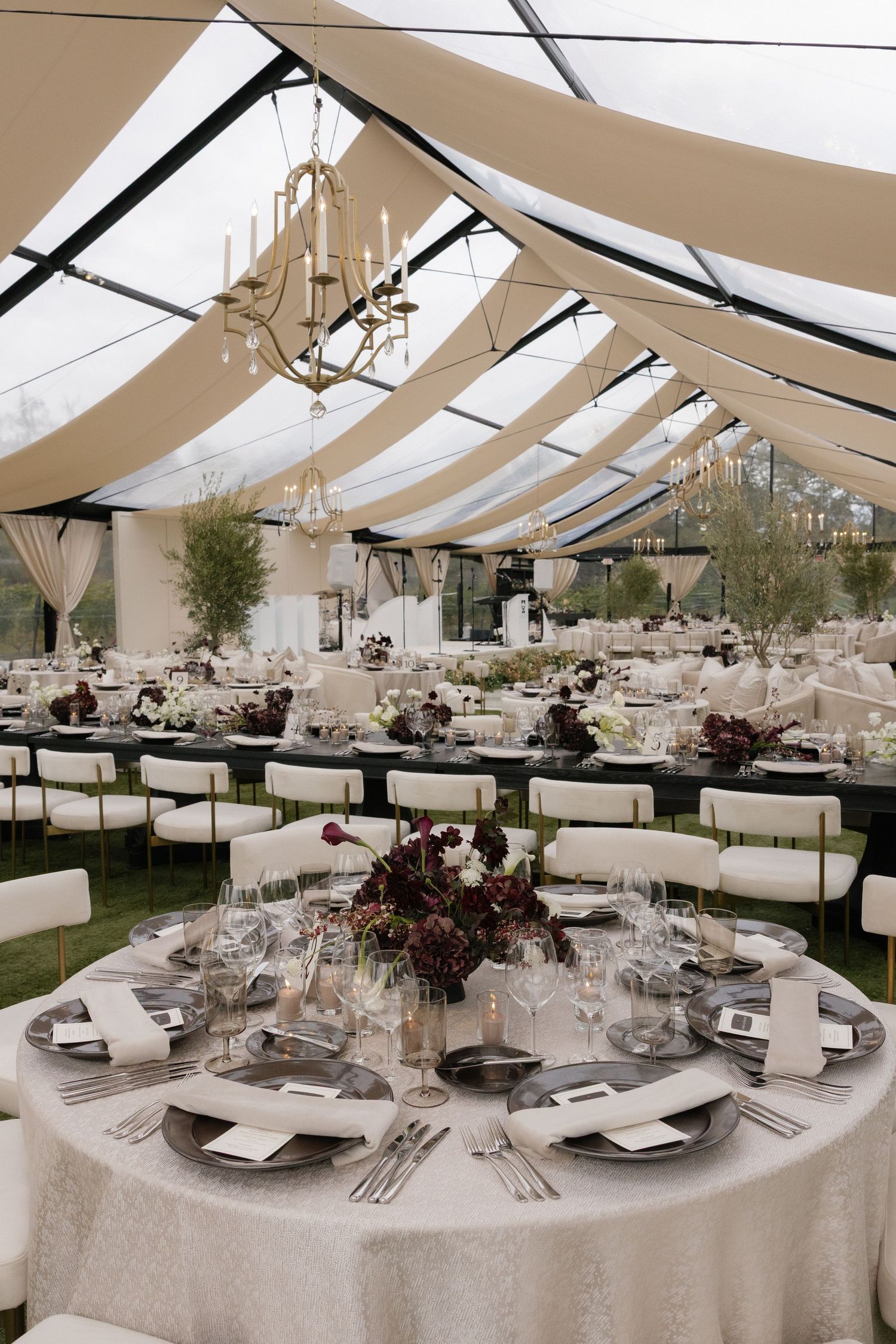 Elegant wedding reception setup with draped ceiling, chandeliers and round tables in white decor.