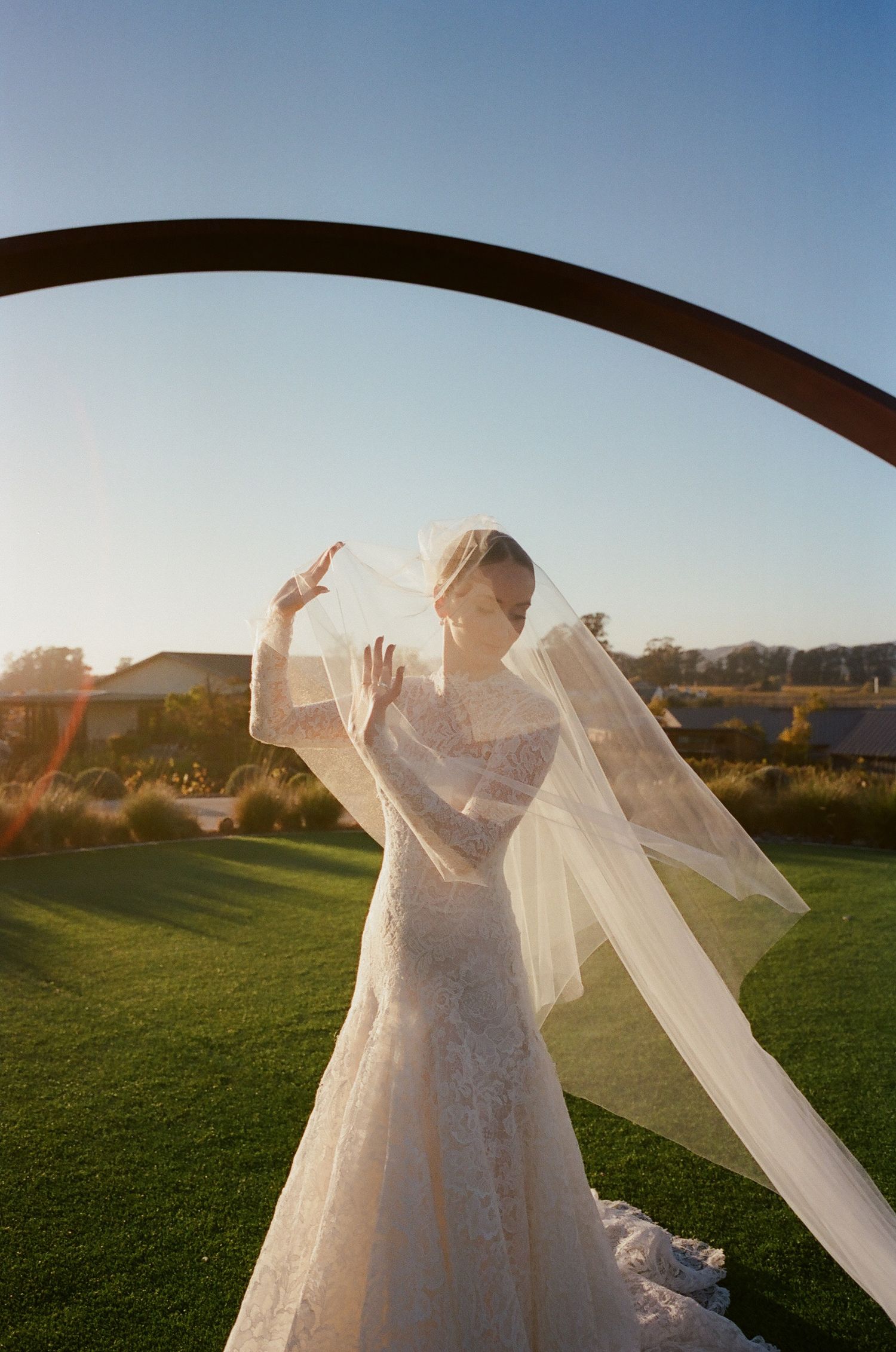 A bride in a lace gown poses playfully with her veil beneath an architectural arch.