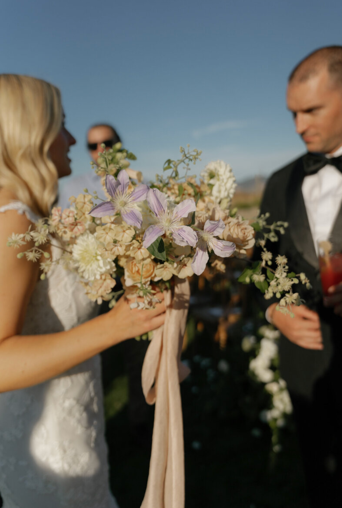 Wedding party holding delicate bouquets with pastel flowers and ribbons against blue sky.