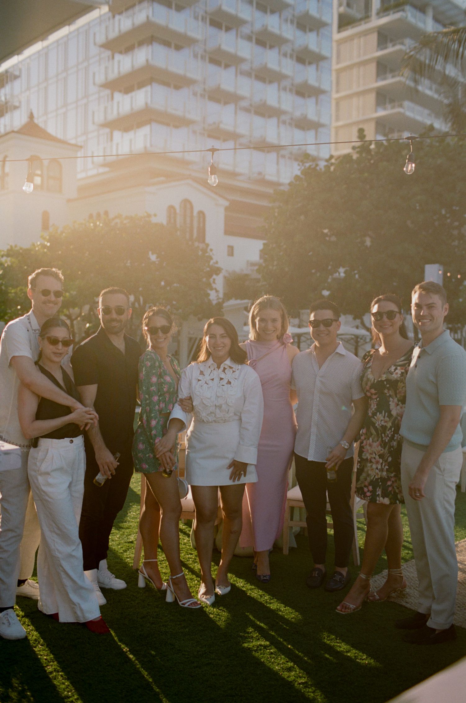 Group photo of wedding guests at sunset with high-rise building in background.