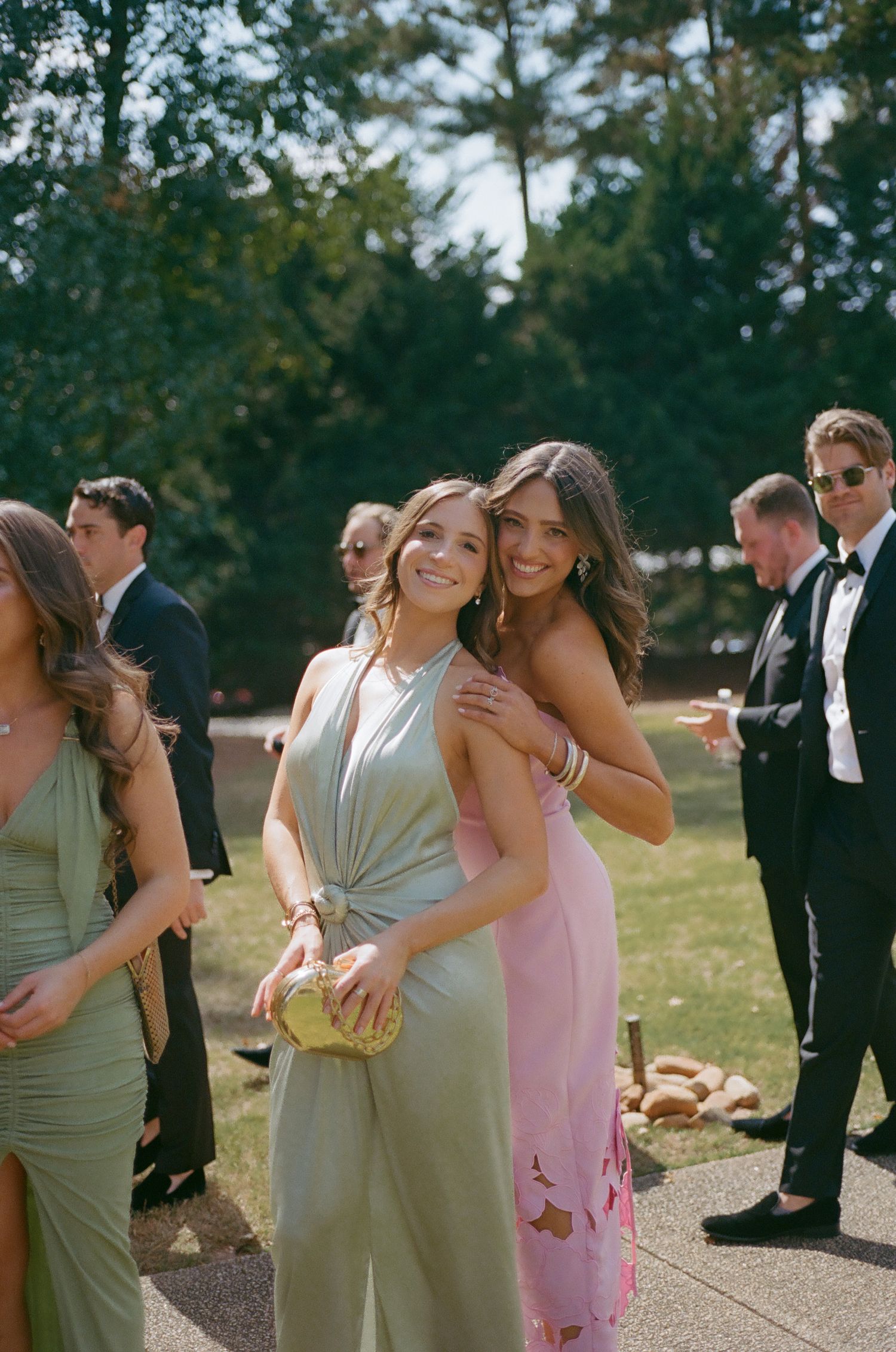 Wedding party members in pink and sage dresses sharing joyful embraces outdoors.