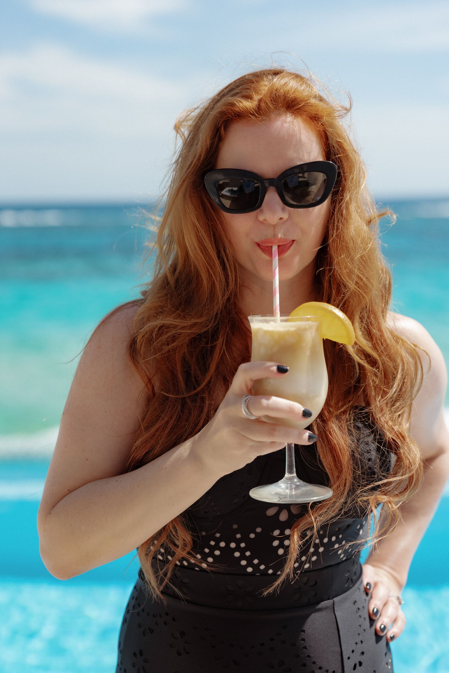 A woman with long red hair wearing sunglasses and drinking a beverage while standing in front of a blue ocean.