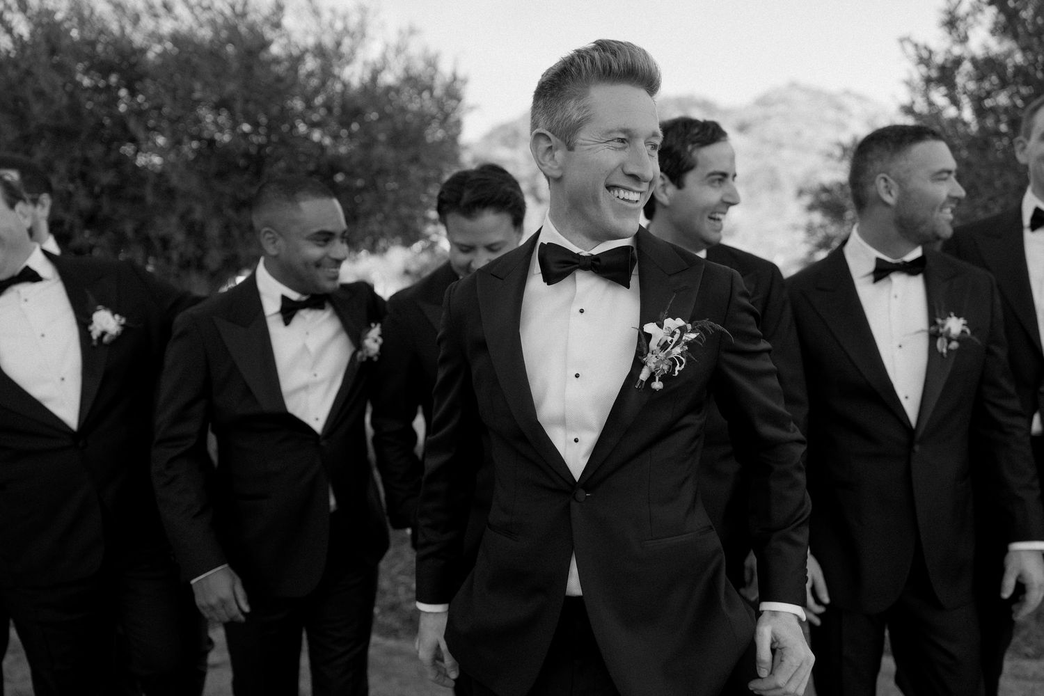 Groomsmen in black tuxedos gather outdoors for wedding photos.