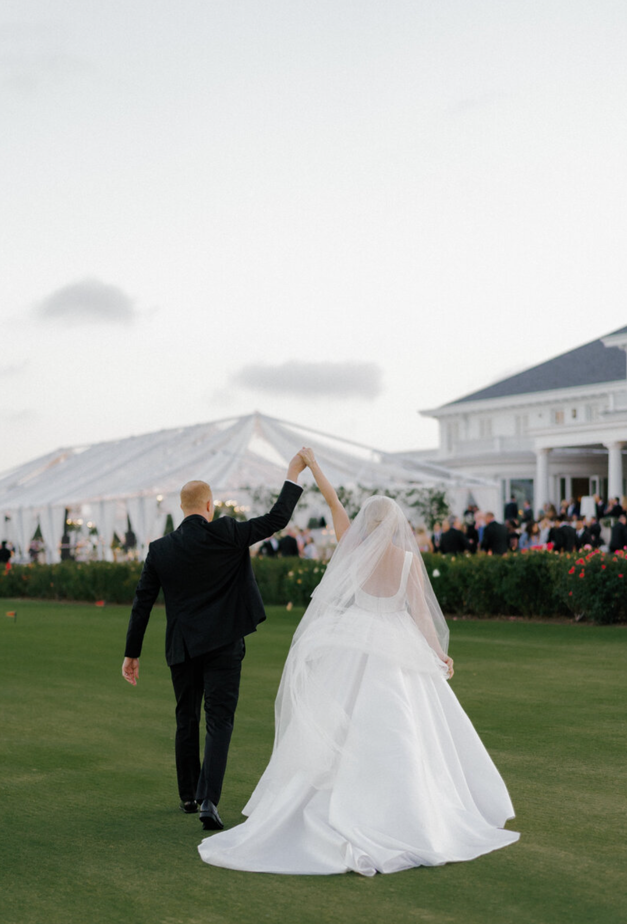 Wedding couple walking and twirling on a lush green lawn with white buildings in the background.