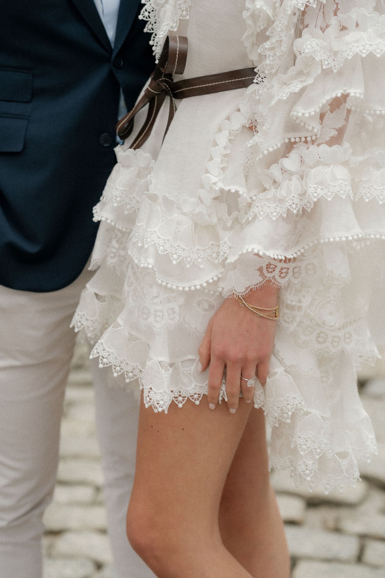 Close-up details of delicate white lace dress and formal attire on cobblestone street.