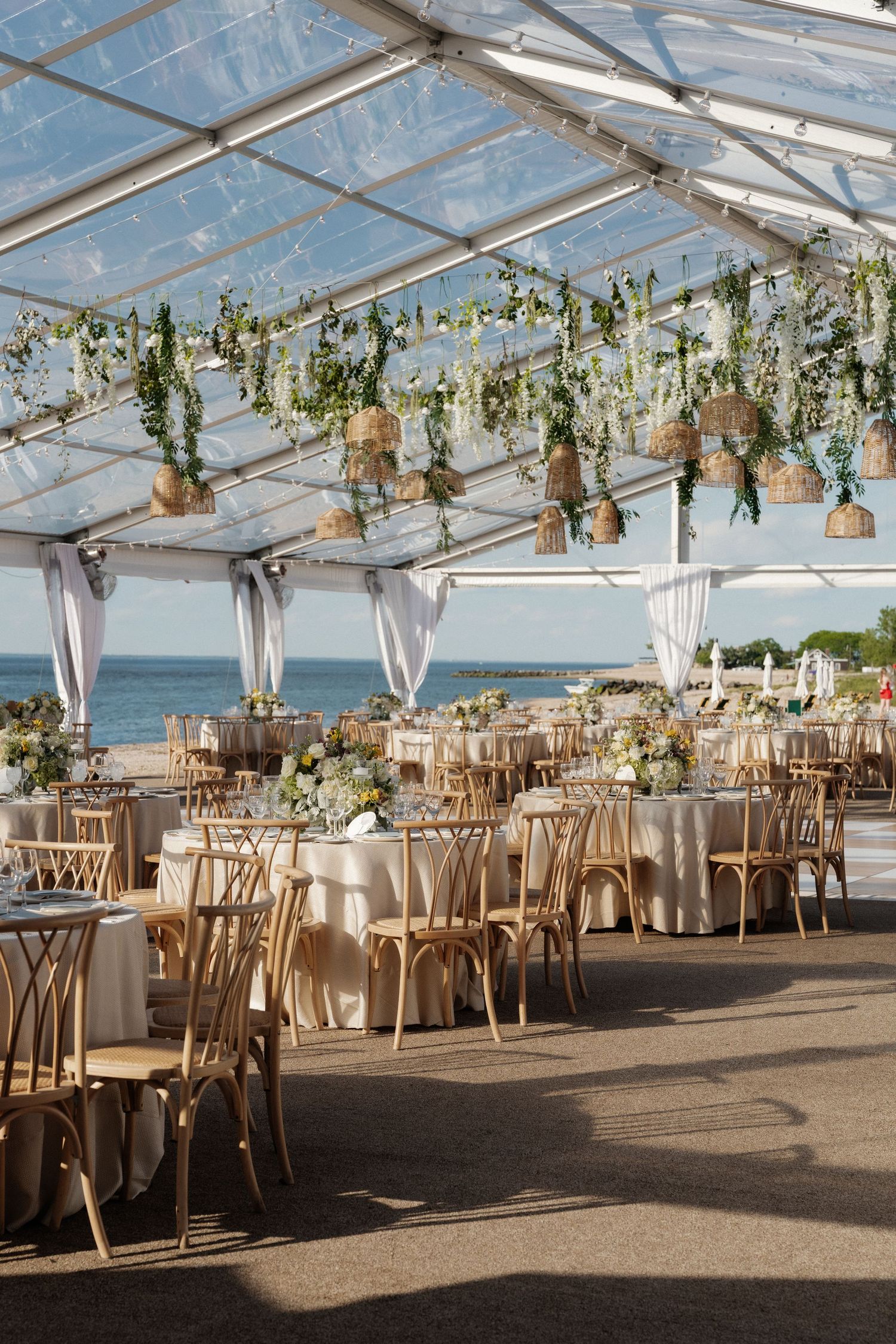 Stunning beachside wedding reception under a clear tent adorned with hanging greenery and wicker lanterns.