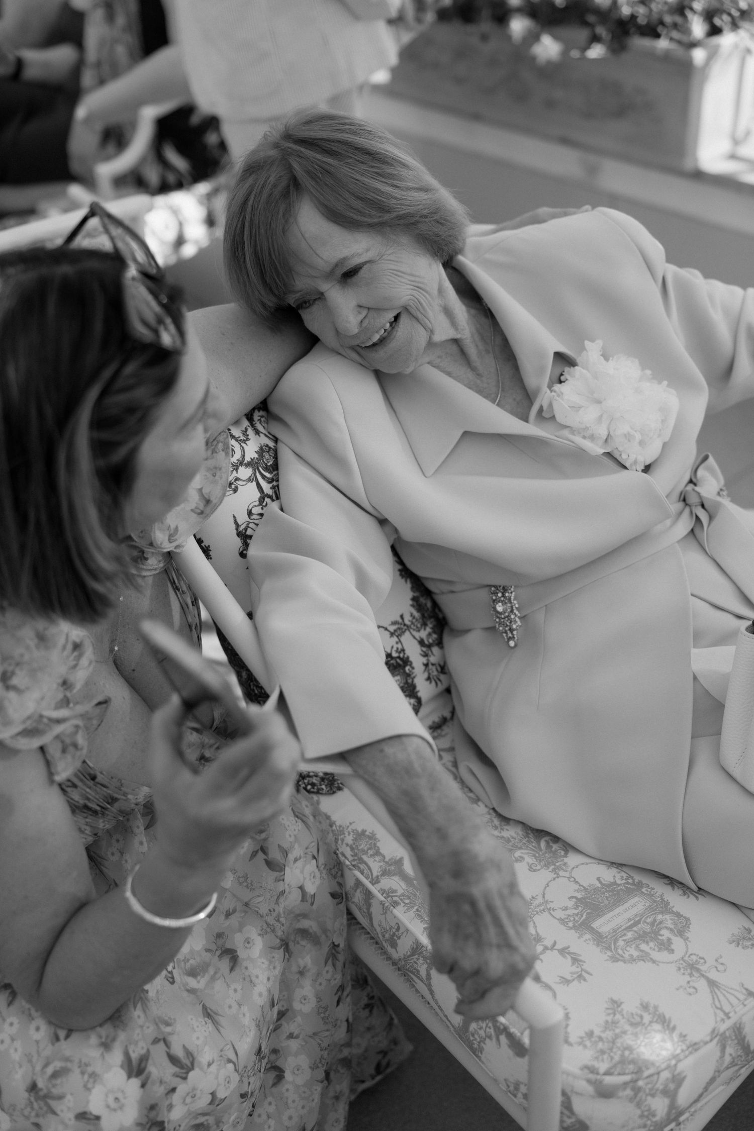 Black and white photo of two women sharing a warm embrace and laugh at an outdoor wedding reception.