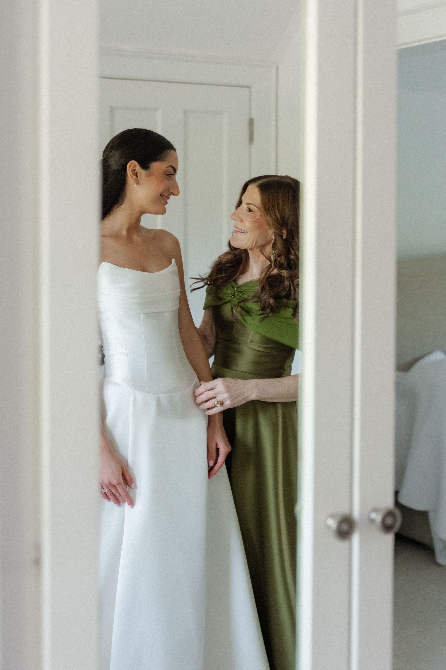 Bride in white strapless gown smiling with bridesmaid in green off-shoulder dress during wedding preparations.
