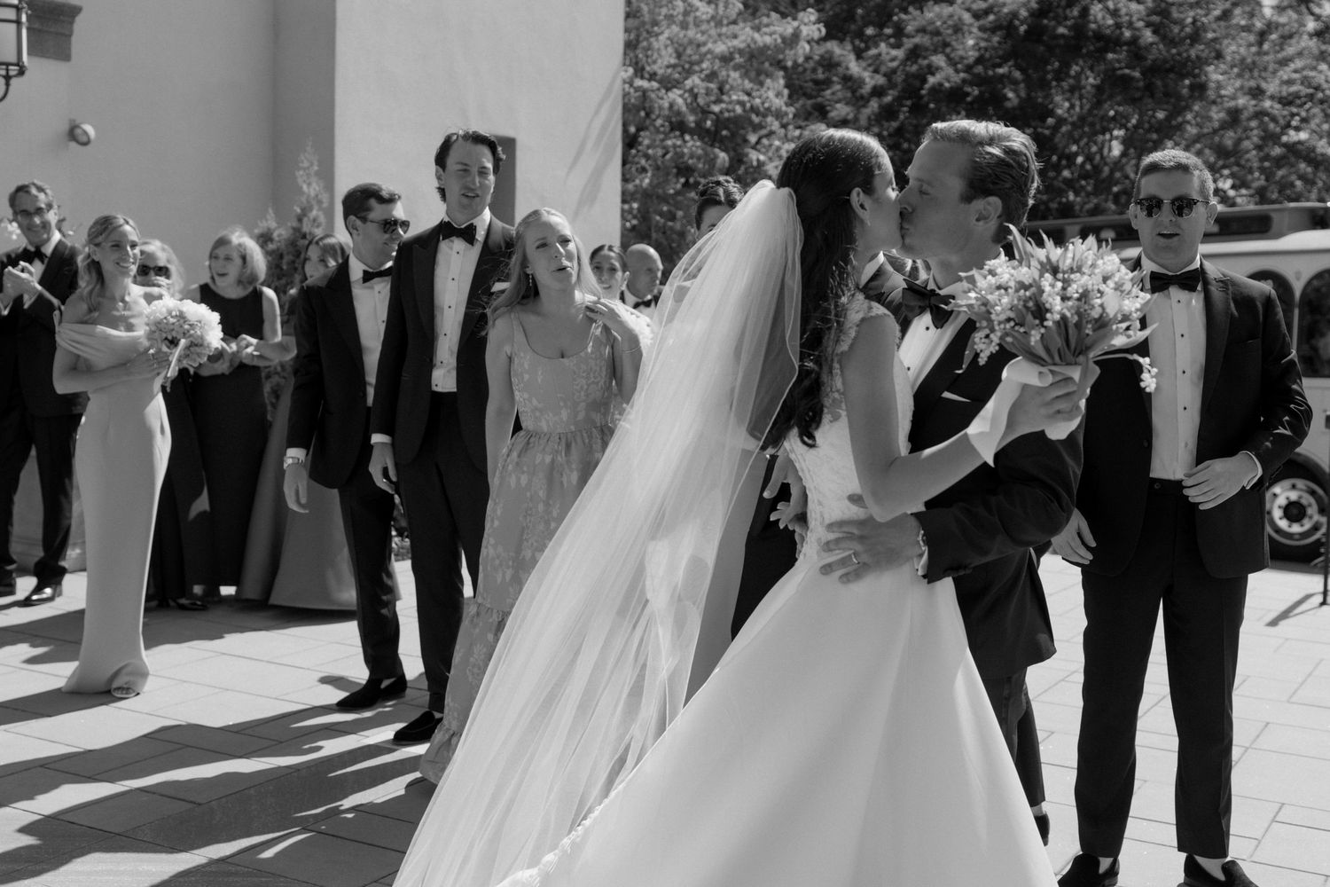 Black and white wedding photo of bride and groom kissing outside church surrounded by guests in formal attire.