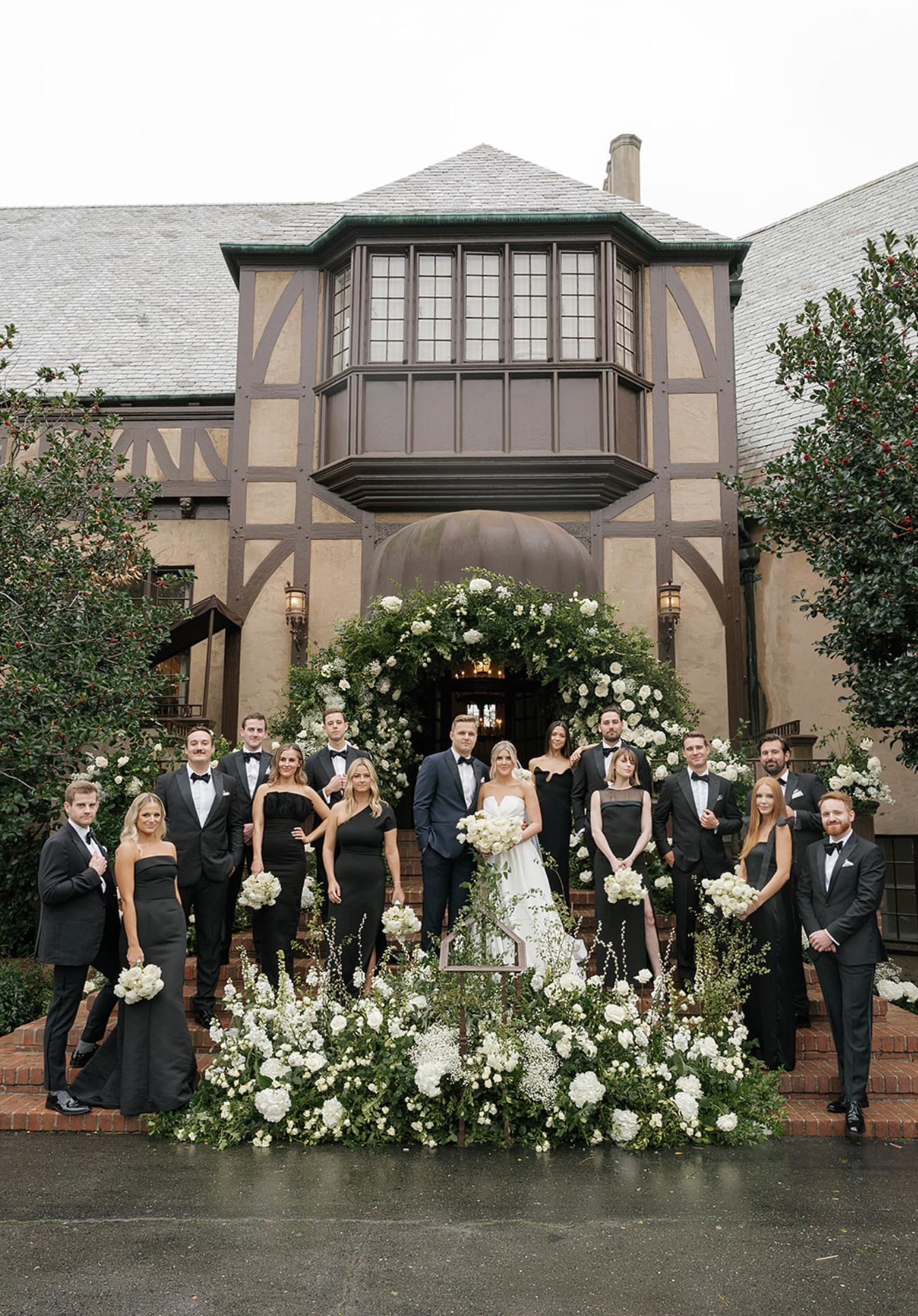 Wedding party in formal attire poses in front of Tudor-style building with floral decorations.