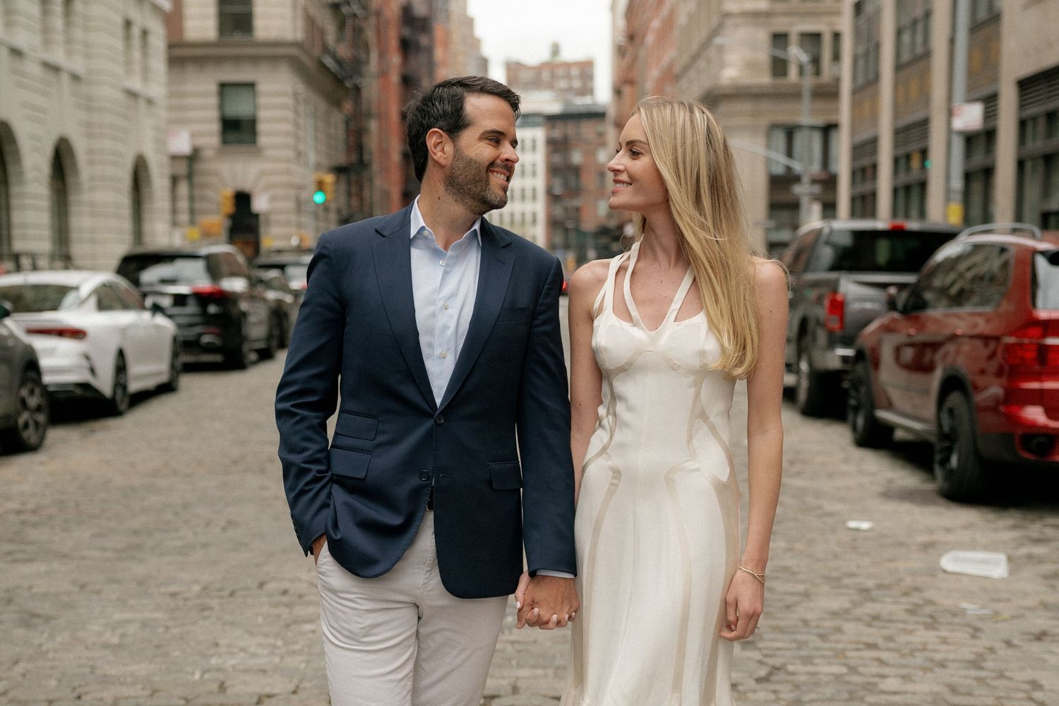 Couple walking together on city street with historic buildings and parked cars in background.