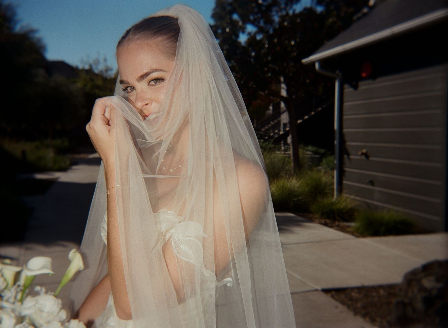 A bride in a flowing veil poses outdoors against a natural backdrop.