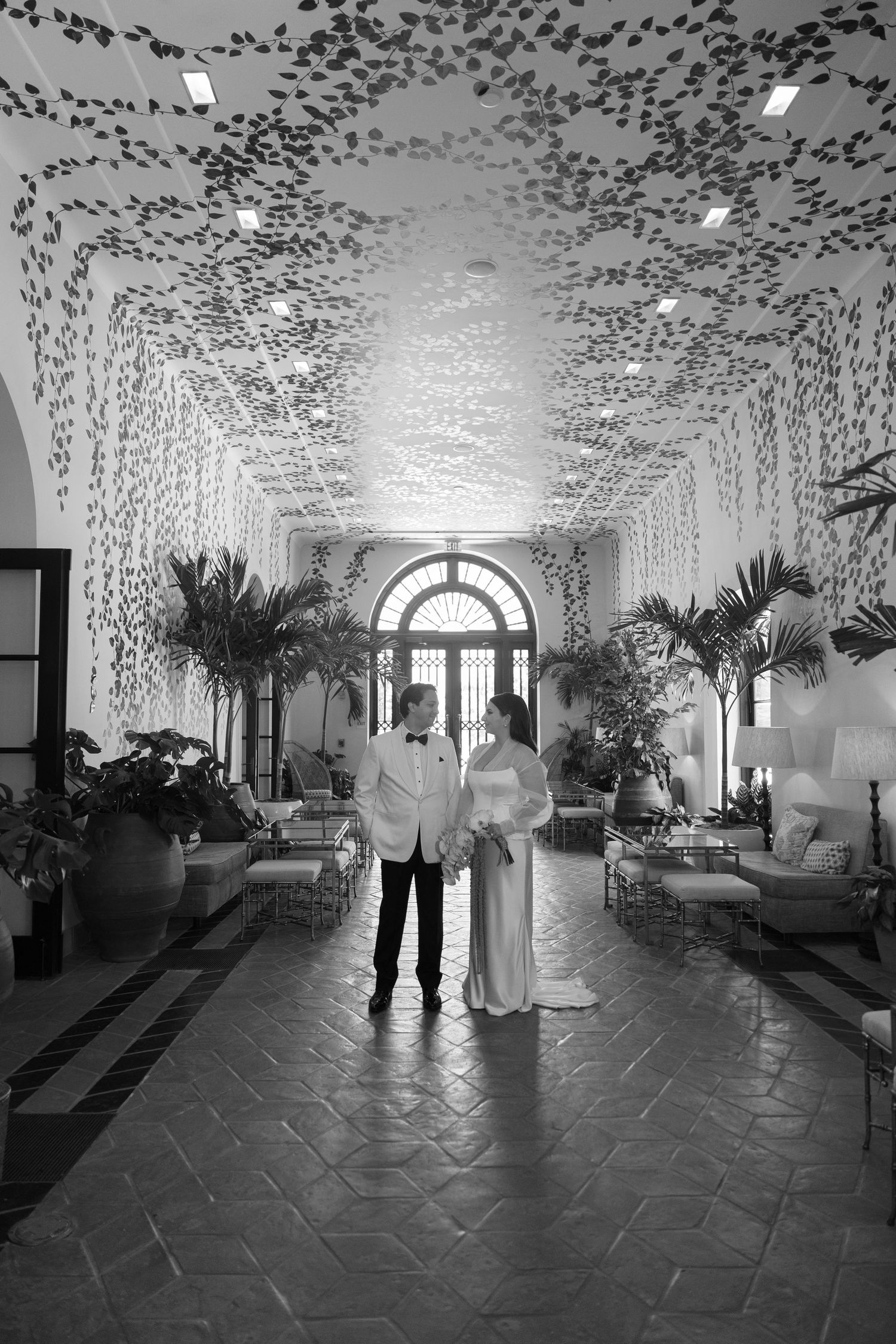 A couple stands under an elegant arched hallway decorated with cascading vines and greenery in black and white.