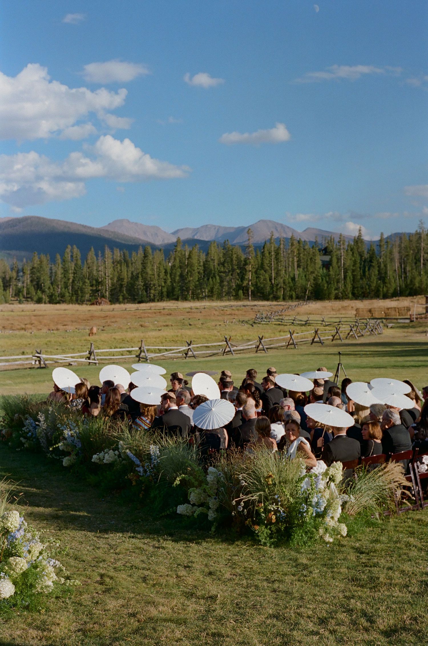 Wedding guests with white parasols gather outdoors against a mountain backdrop on a sunny day.