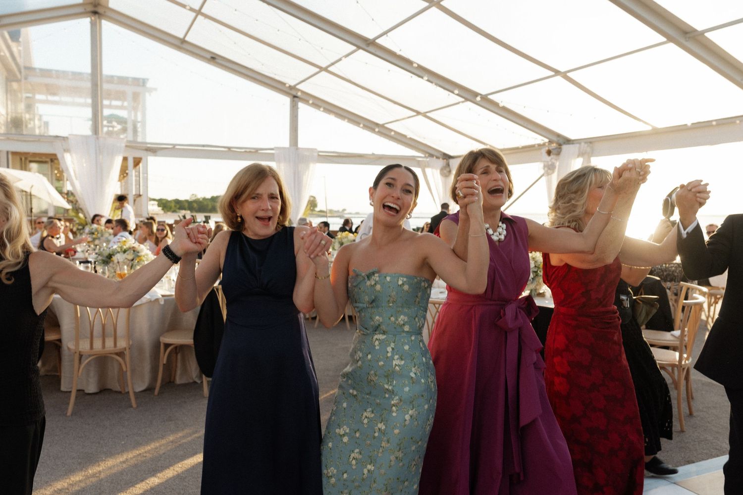 Four women in colorful formal gowns dancing joyfully together at an elegant tented wedding reception.