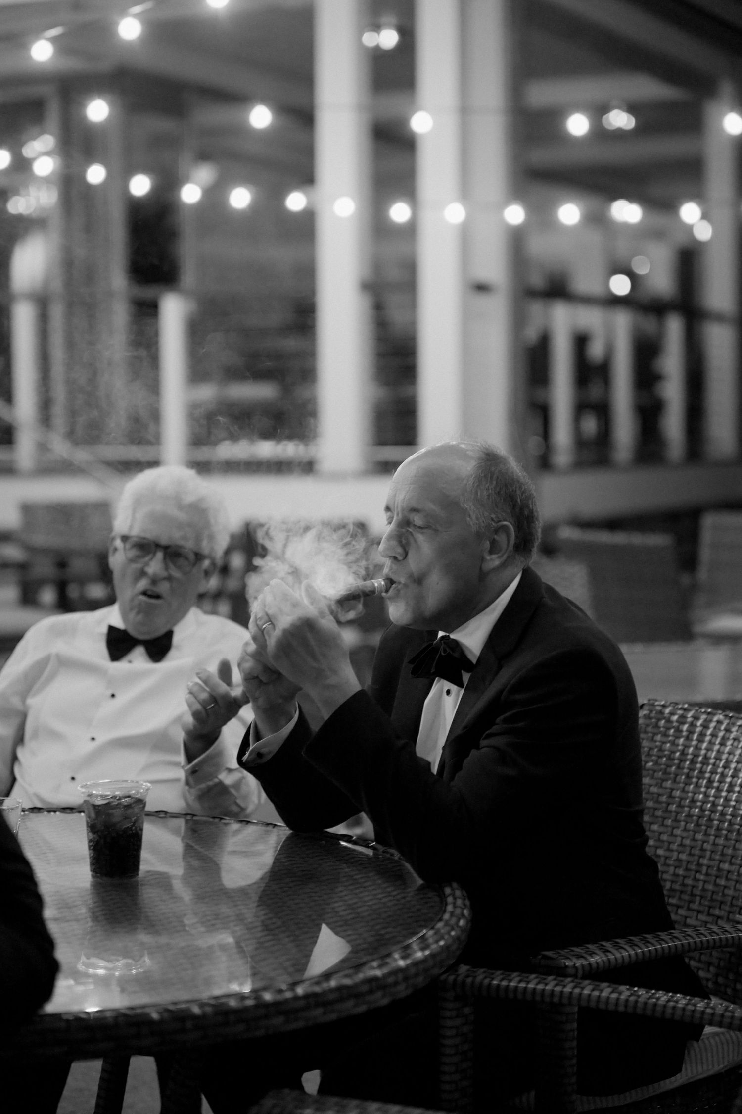 Elegant older gentleman in tuxedo smoking cigar at outdoor wedding reception under string lights.