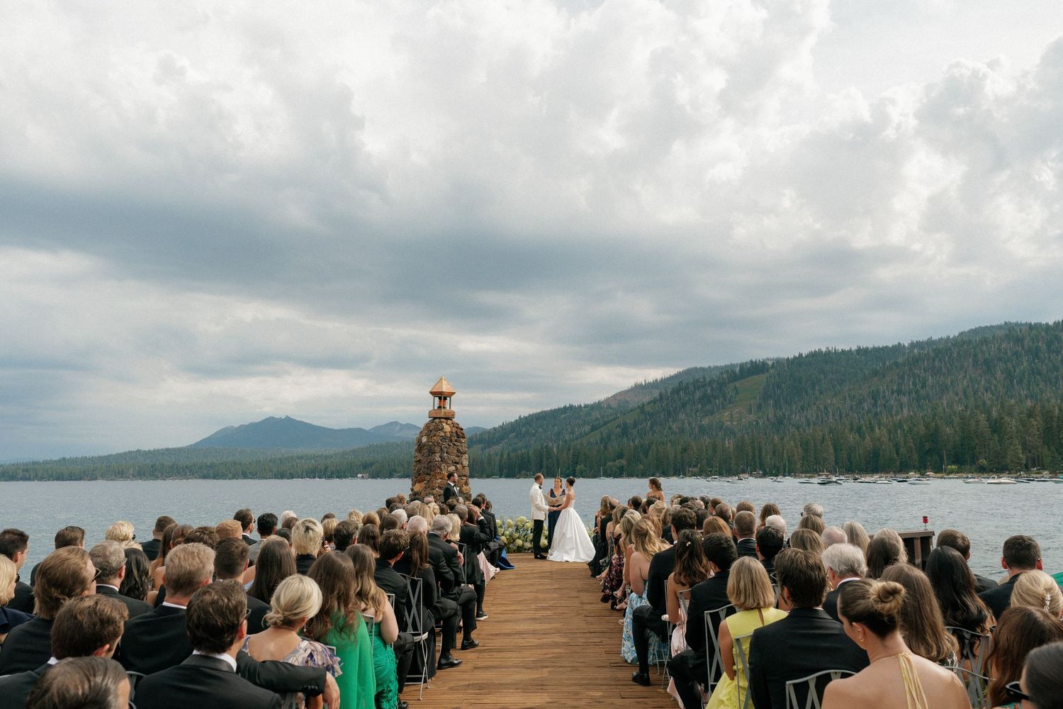 Outdoor wedding ceremony overlooking mountain lake with guests seated on wooden chairs.