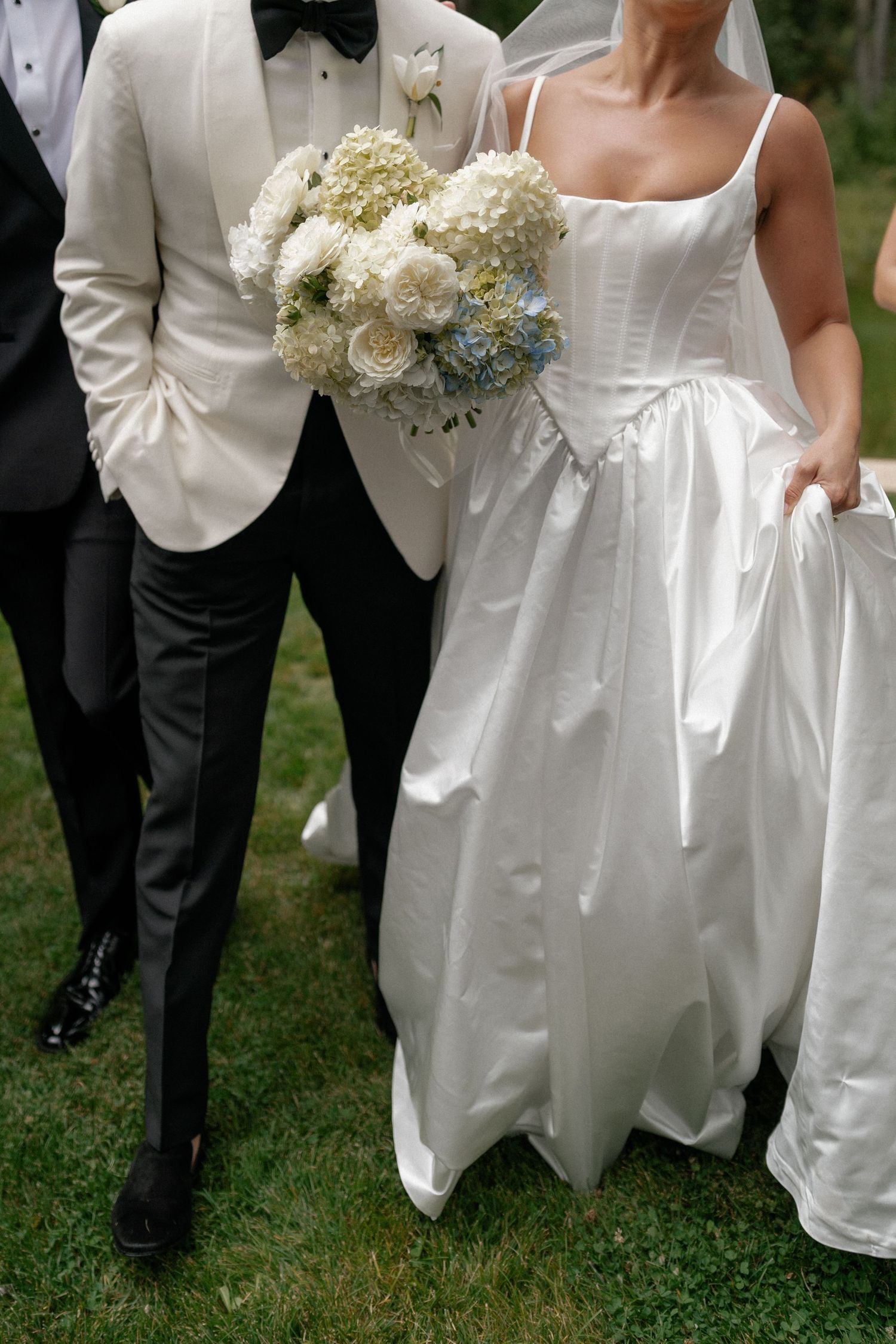 Wedding party members in white dresses and tuxedos gather on grass holding bouquets.