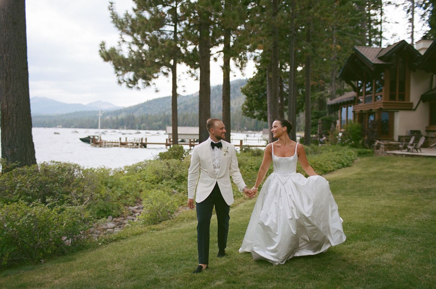 A couple walks along a scenic lakeside path with mountains and pine trees in the background.