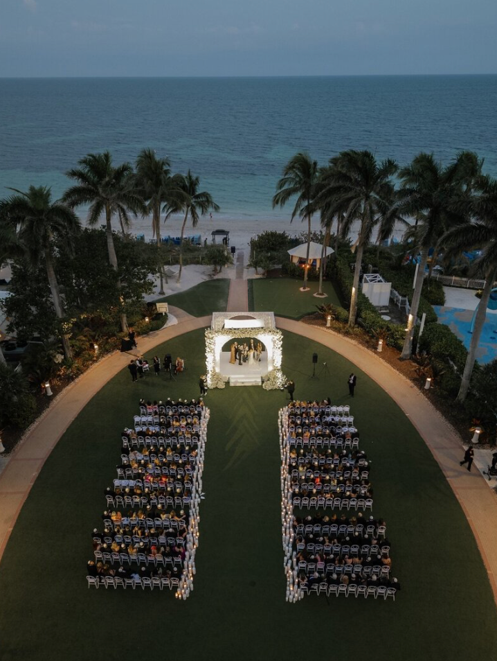 Aerial view of an outdoor wedding ceremony setup with white chairs and gazebo surrounded by palm trees overlooking the ocean.