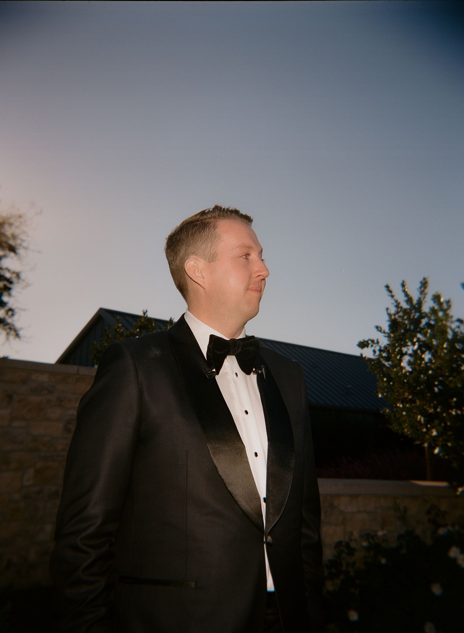 A groom in a black tuxedo and white bow tie stands outside at dusk in a series of artistic photos.