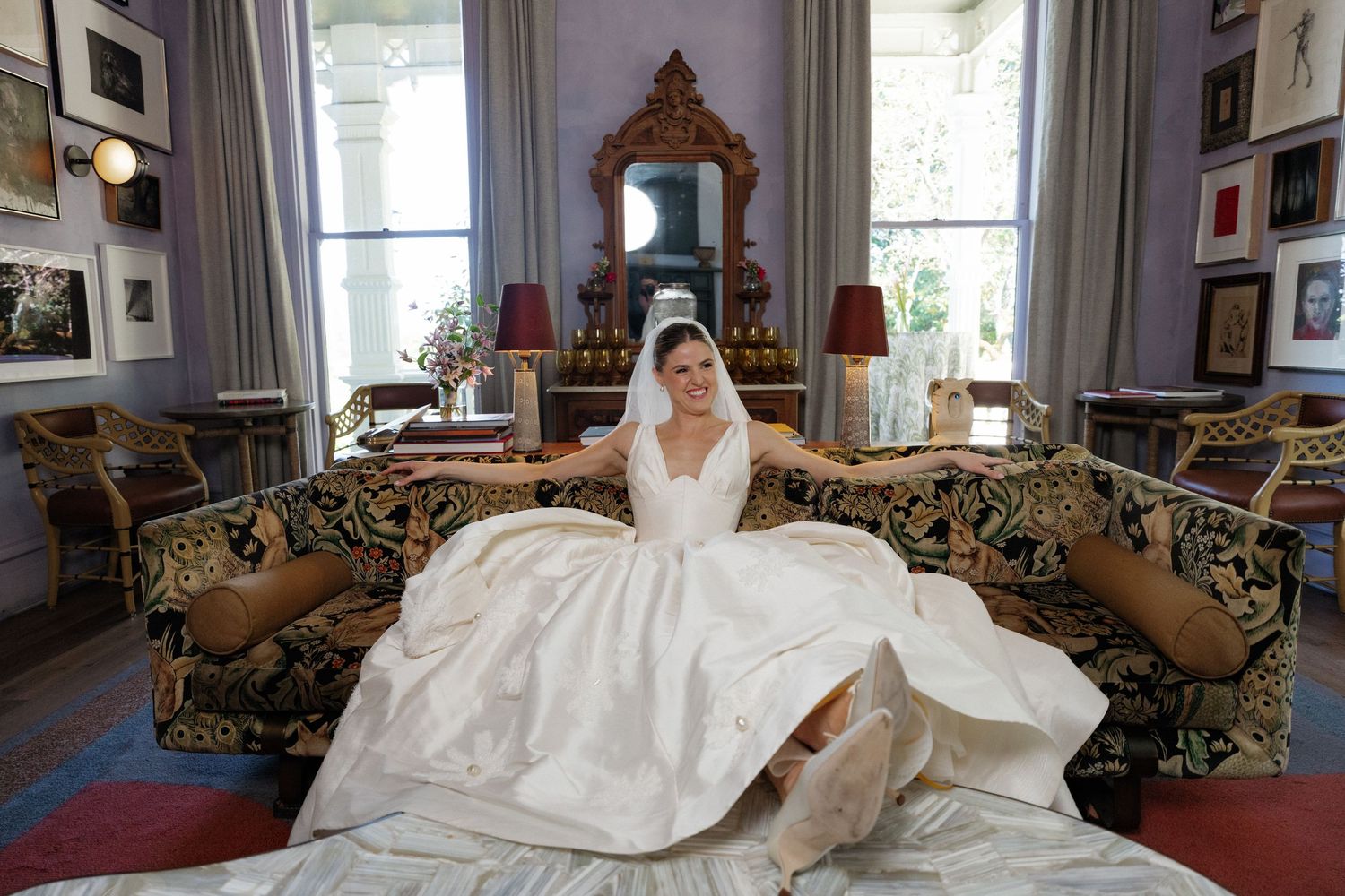 Bride in a voluminous white wedding gown sitting on a floral sofa in an elegantly decorated room with artwork on walls.