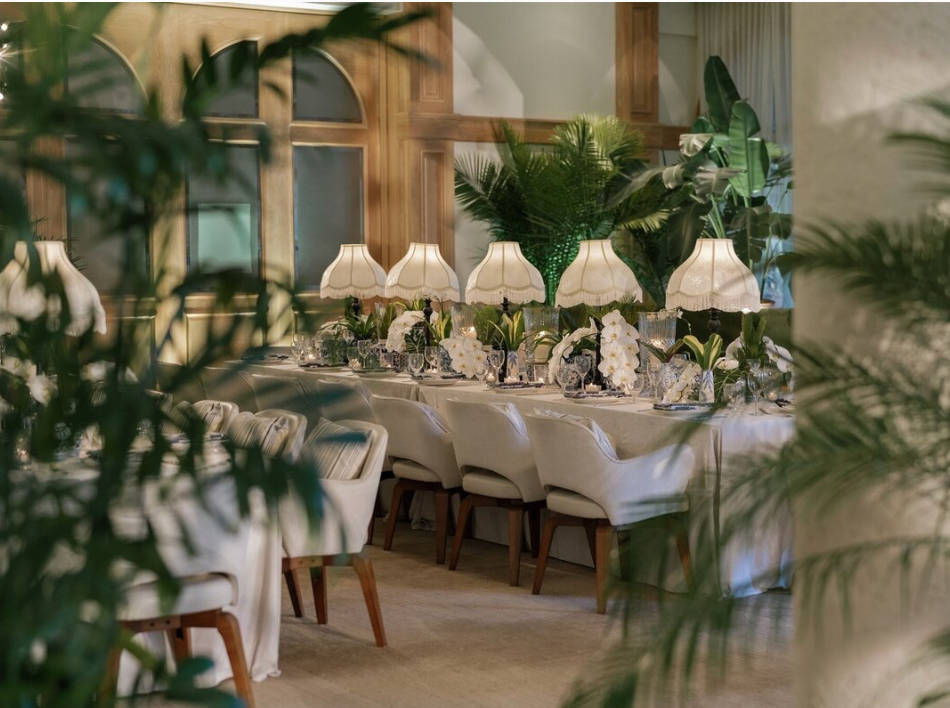 Elegant wedding reception table setting with white linens and floral centerpieces framed by tropical plants.