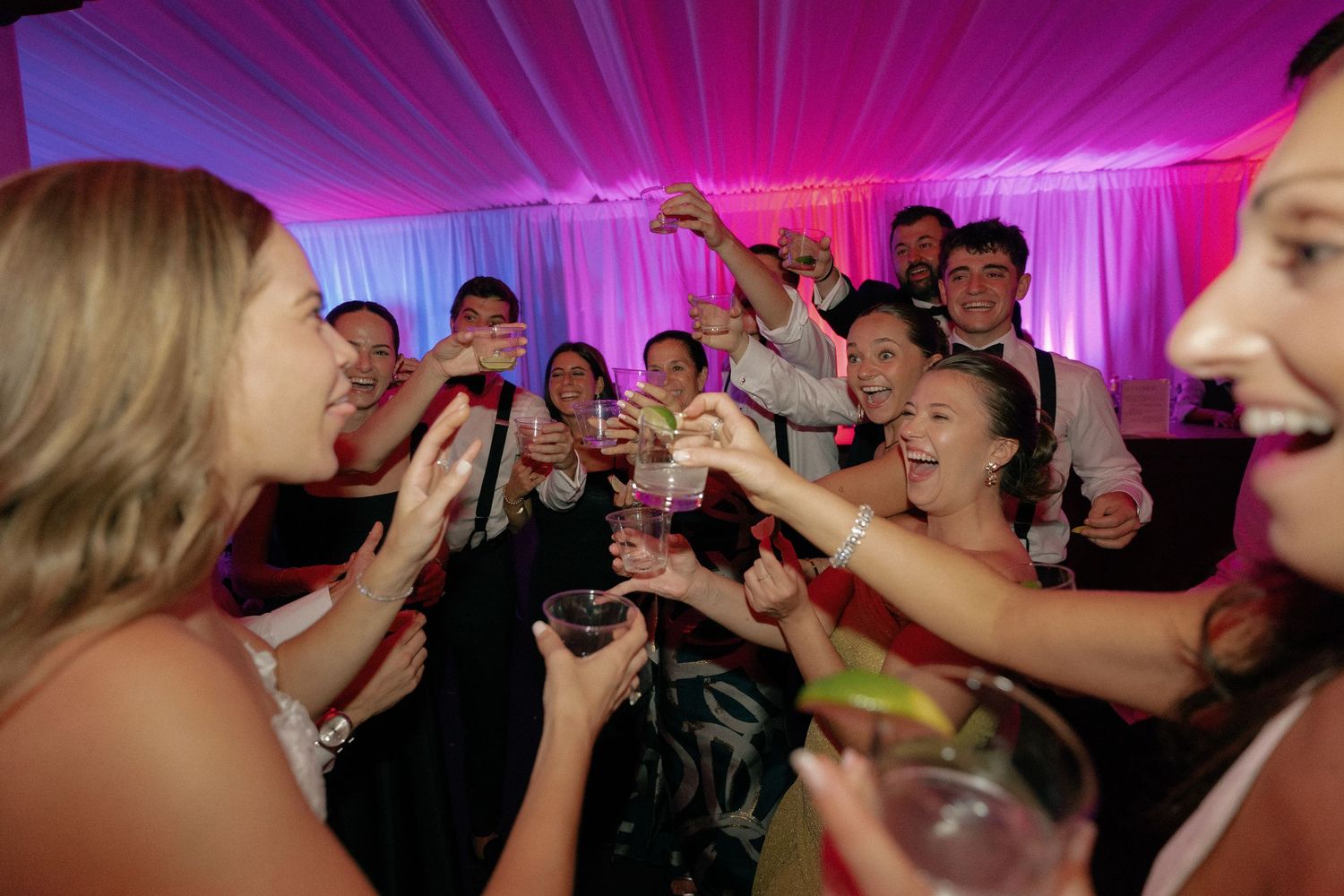 Wedding party dancing under vibrant purple lighting in an elegant tent venue.