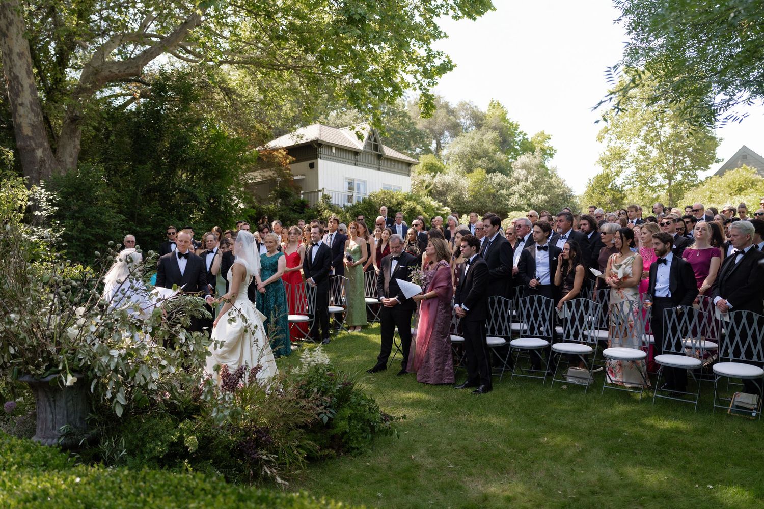 Outdoor wedding ceremony on a sunny day with guests seated on chairs in a lush garden as the bride walks down the aisle.