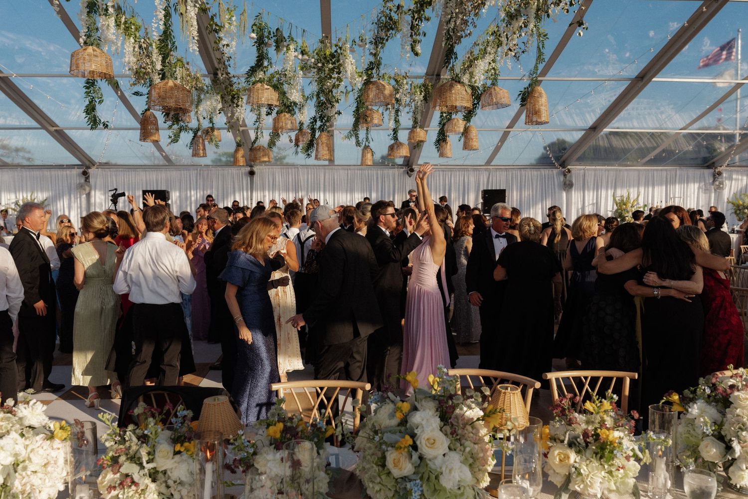 Guests dancing under floral decorations in a glass marquee at an elegant wedding reception.