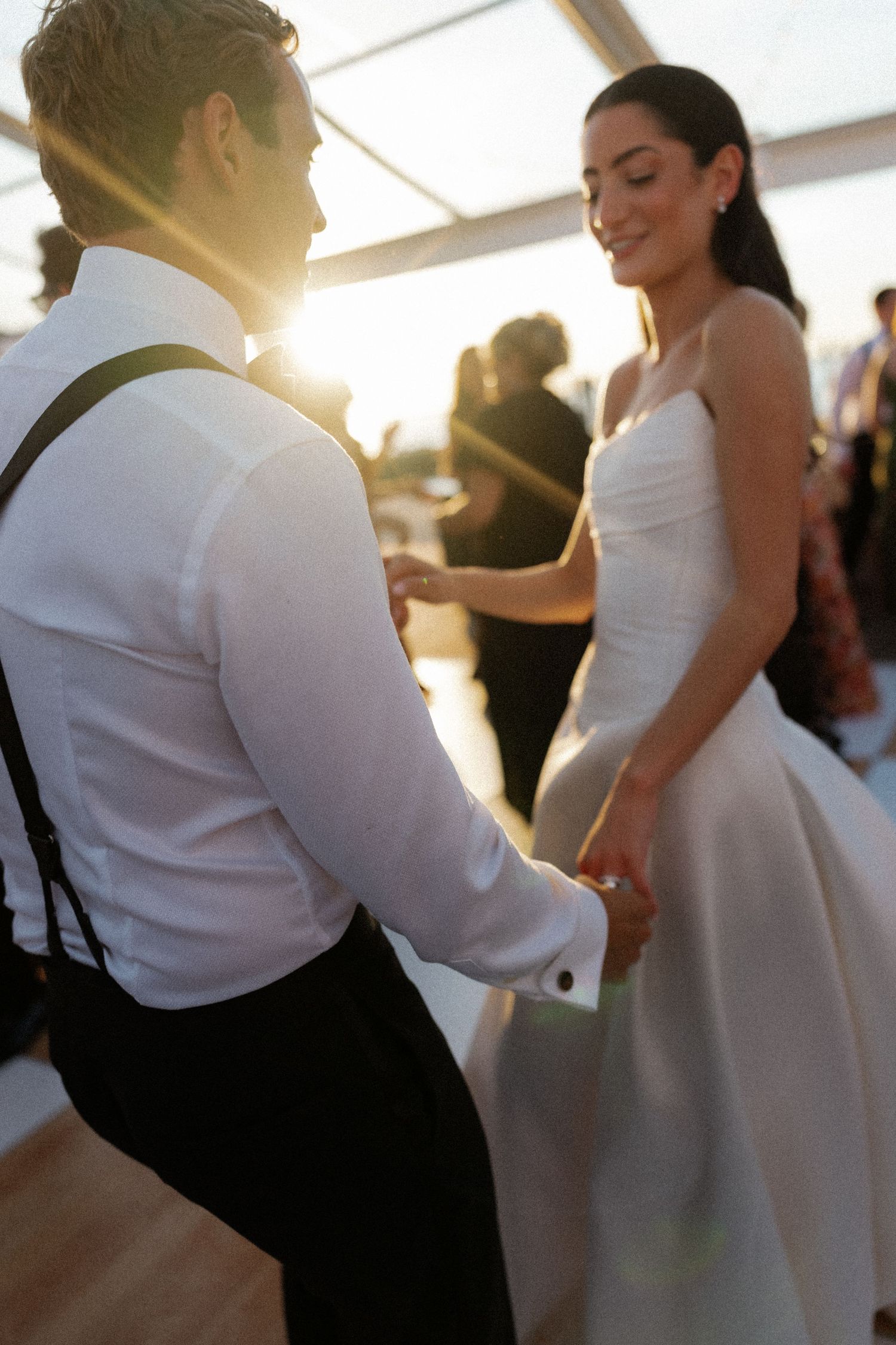 Bride and groom sharing first dance bathed in golden sunset light, bride in white gown, groom in suspenders.