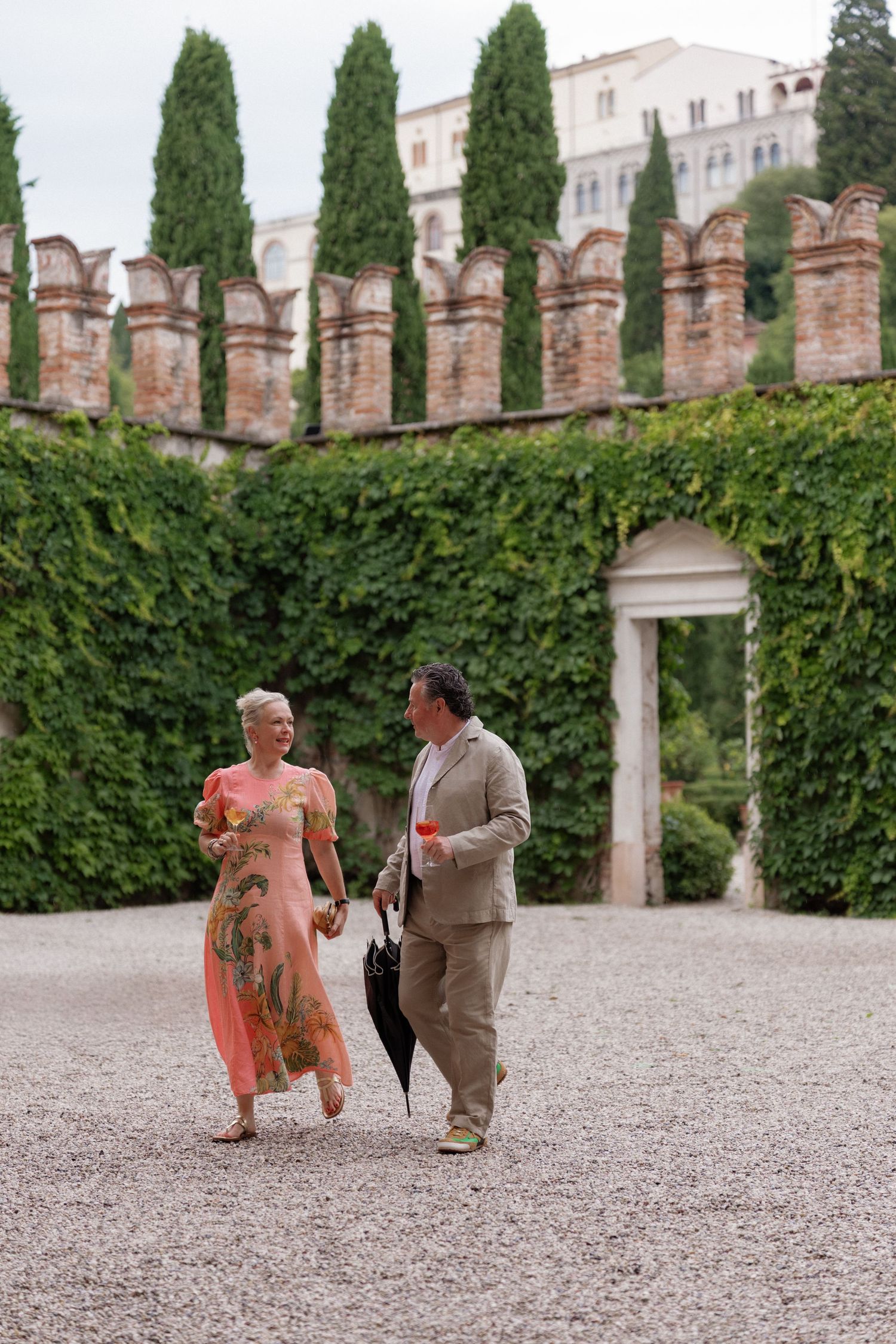 Elegant couple strolling through an ivy-covered Italian garden with cypress trees and historic stone walls.