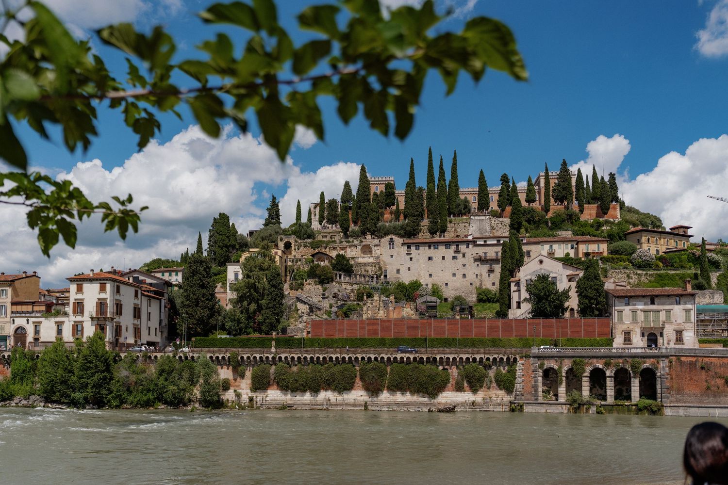 Historic Verona cityscape with cypress trees atop a hill overlooking the Adige River under a partly cloudy sky.