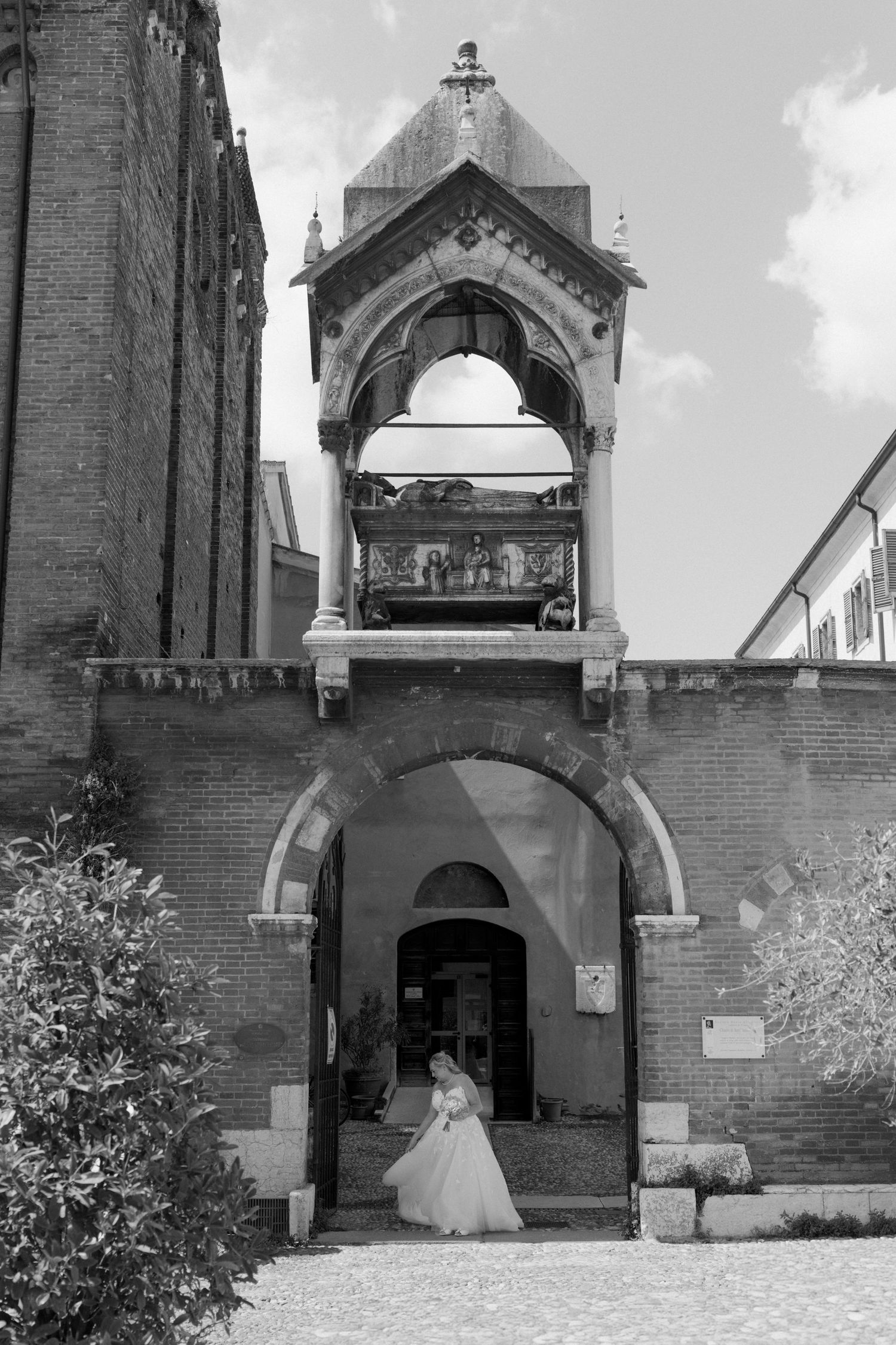 Black and white photo of a bride in wedding dress standing under ornate brick archway of historic Italian church.